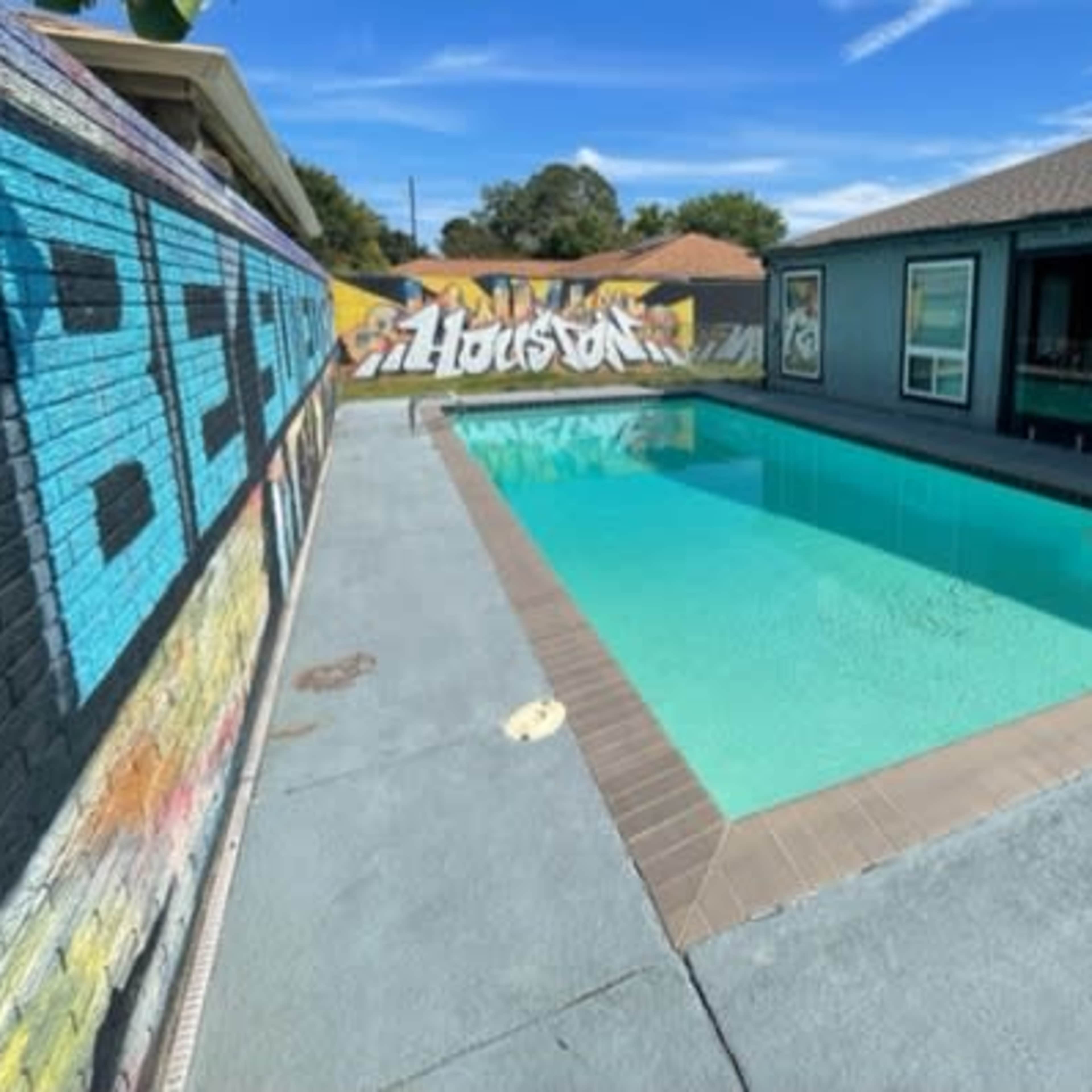 A clear swimming pool is surrounded by a concrete deck and a colorful mural on one side of the yard.
