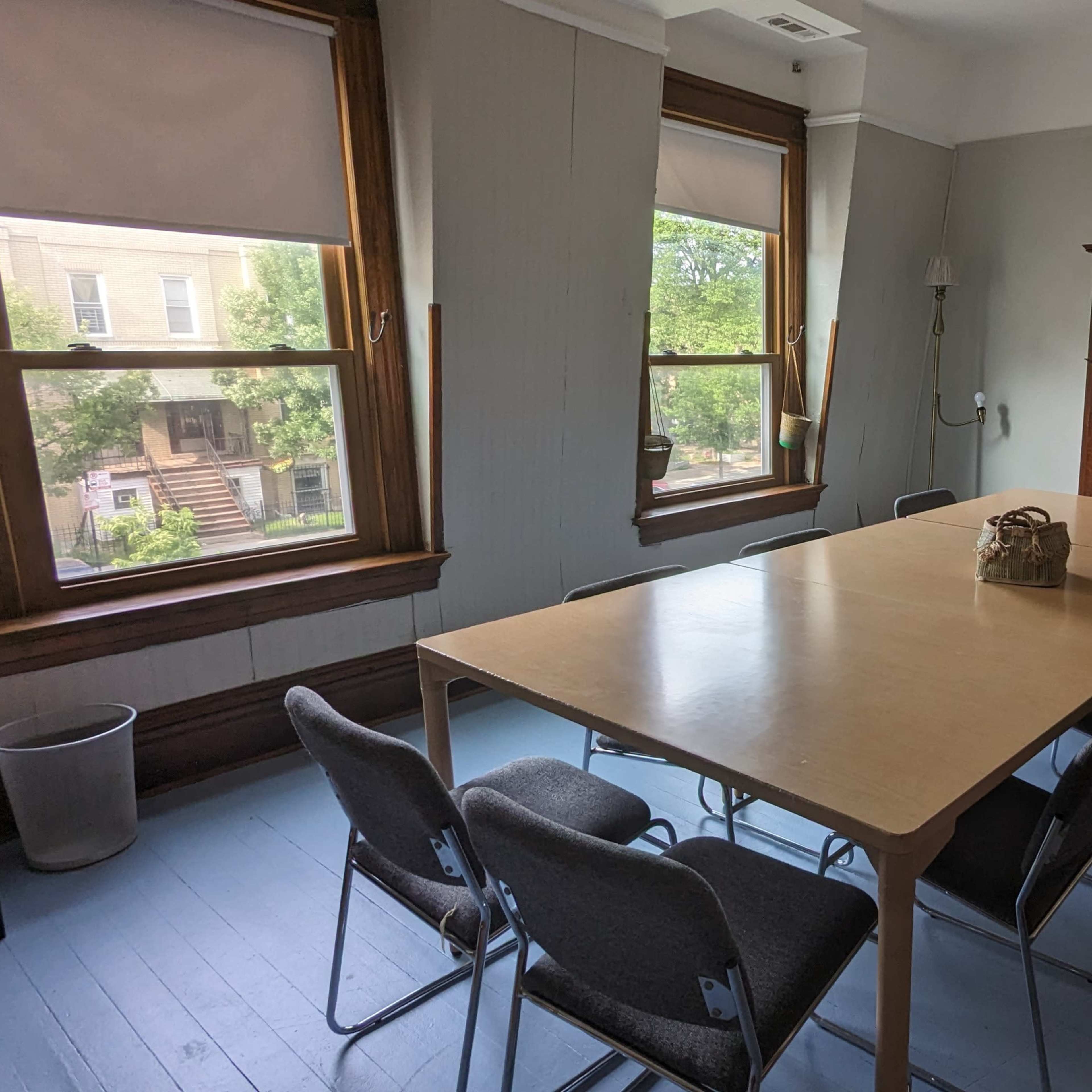A meeting room with a large wooden table surrounded by chairs, two windows providing natural light, and a decorative fireplace.