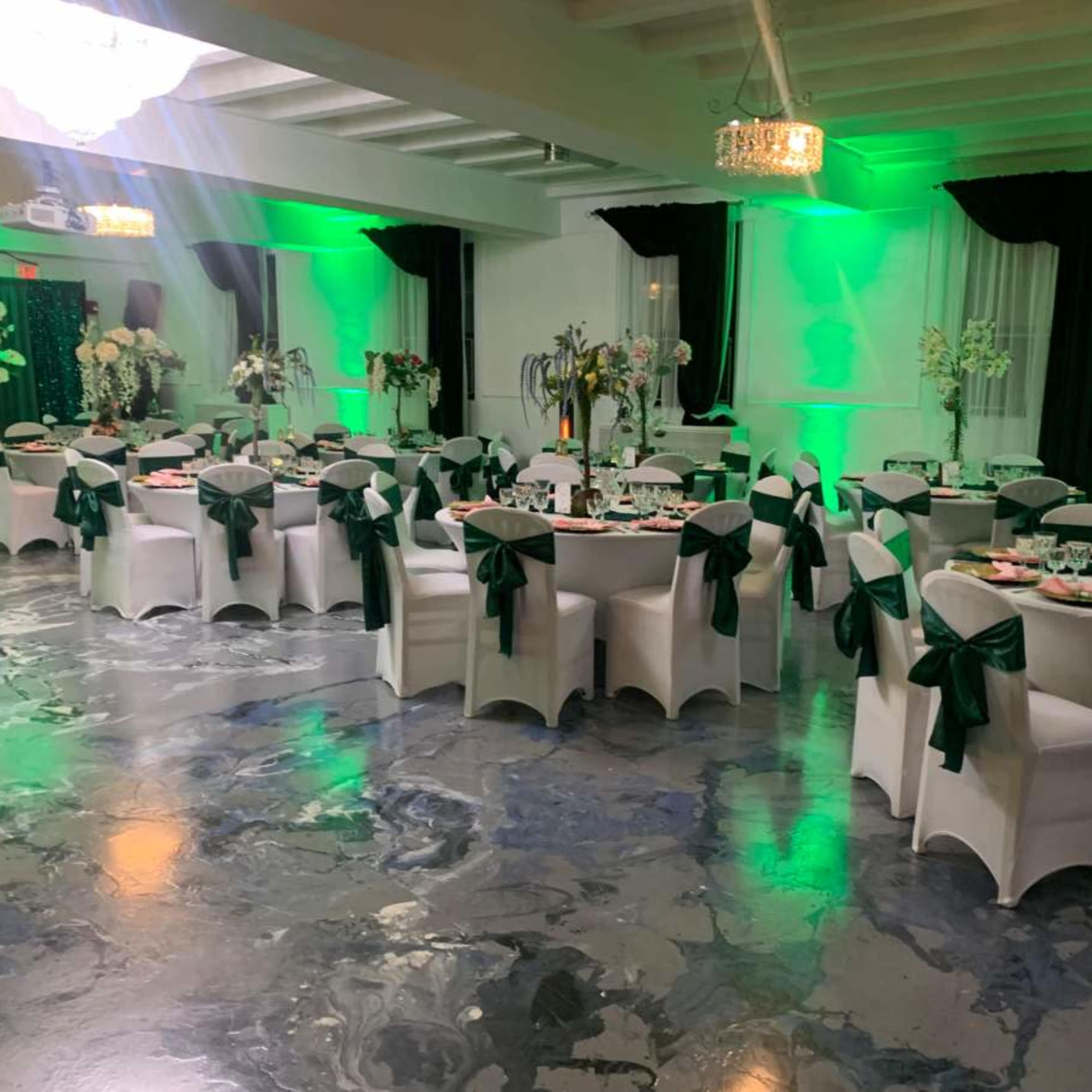 A banquet hall is set up with round tables dressed in white linens and green bows, surrounded by floral centerpieces and illuminated by green lighting.