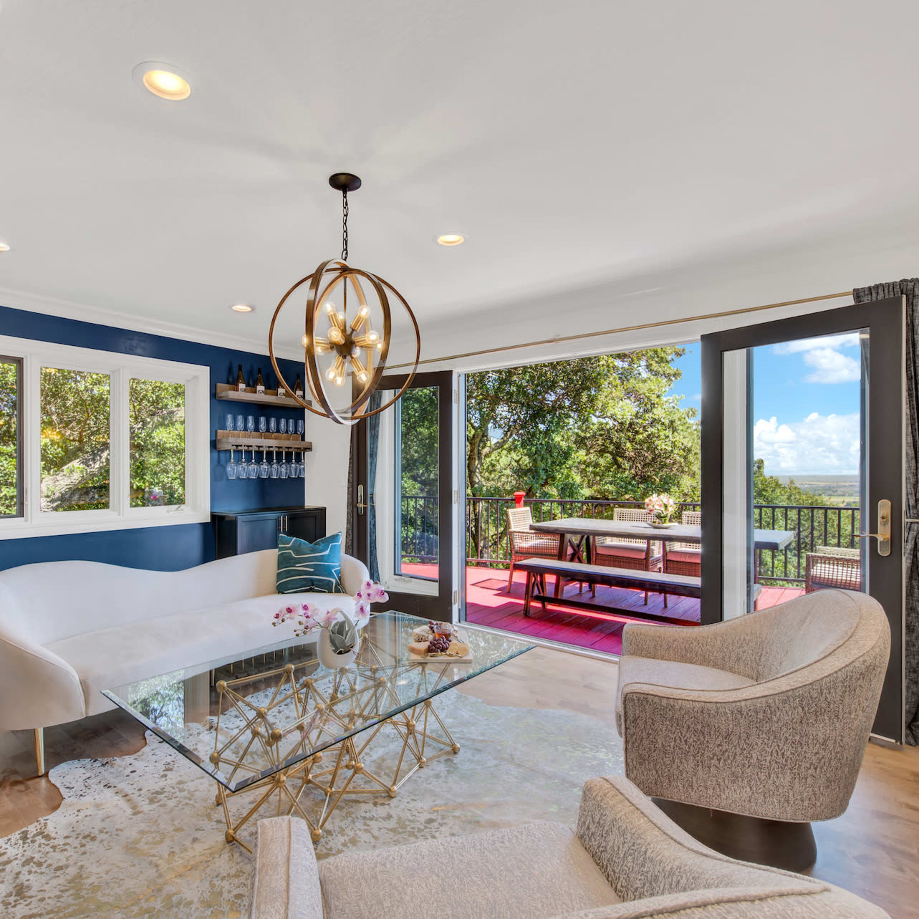 A modern living room features a glass coffee table, a white curved sofa, and large windows that open to an outdoor patio with a dining area and scenic views.