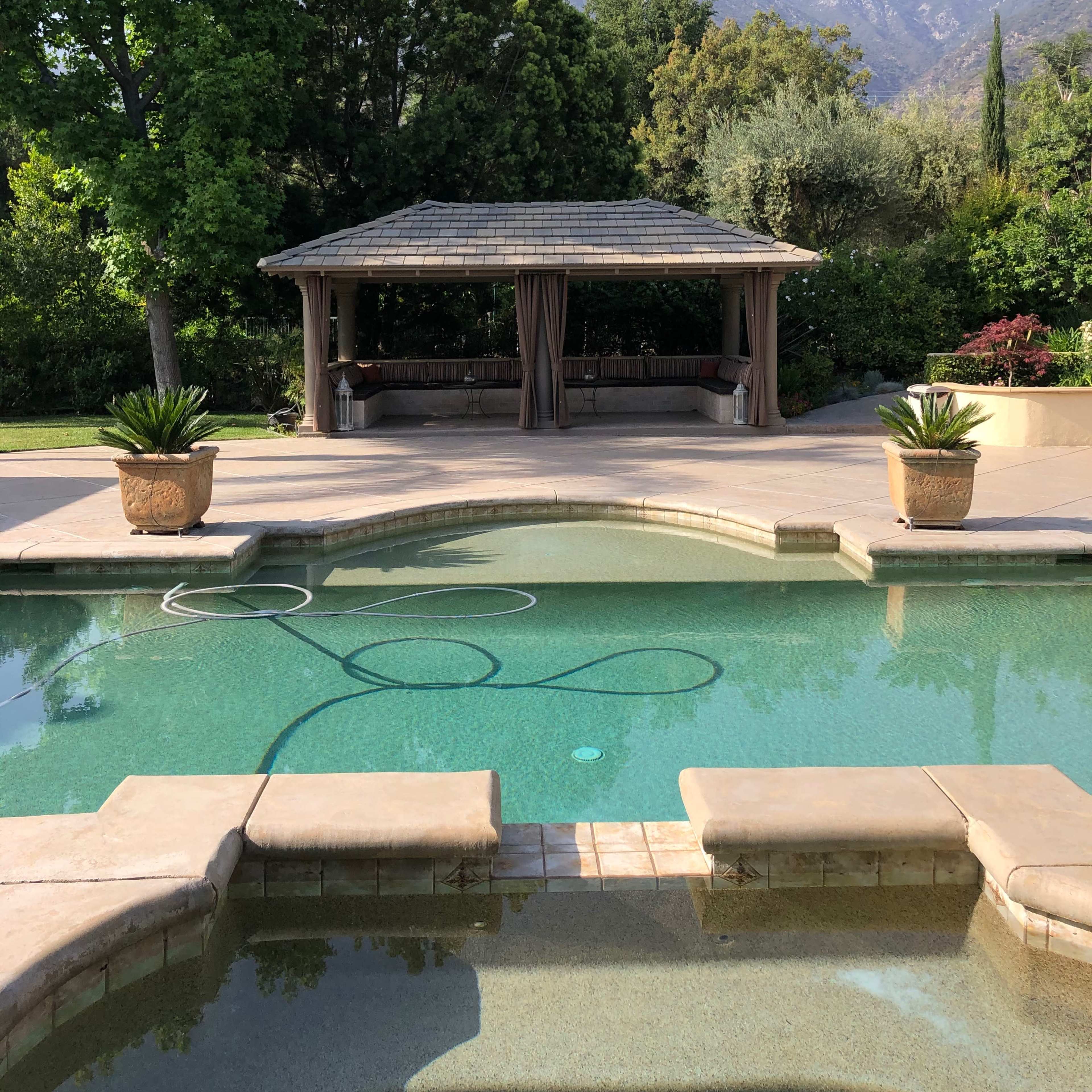 A swimming pool with a stone border reflects a gazebo surrounded by greenery and mountains in the background.