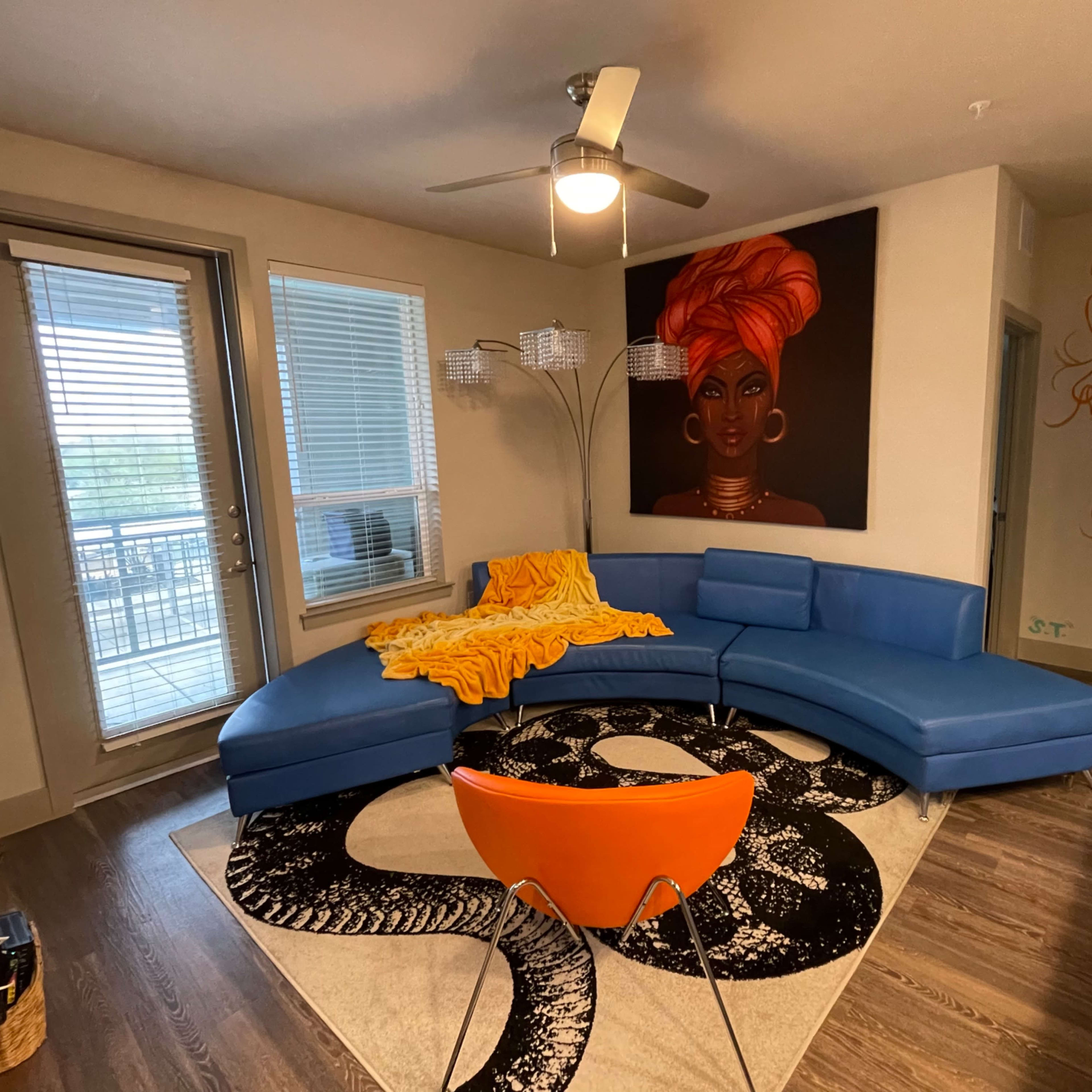 The scene shows a modern living room featuring a blue sectional sofa, an orange chair, a large painting of a woman on the wall, and a patterned rug on a wooden floor.
