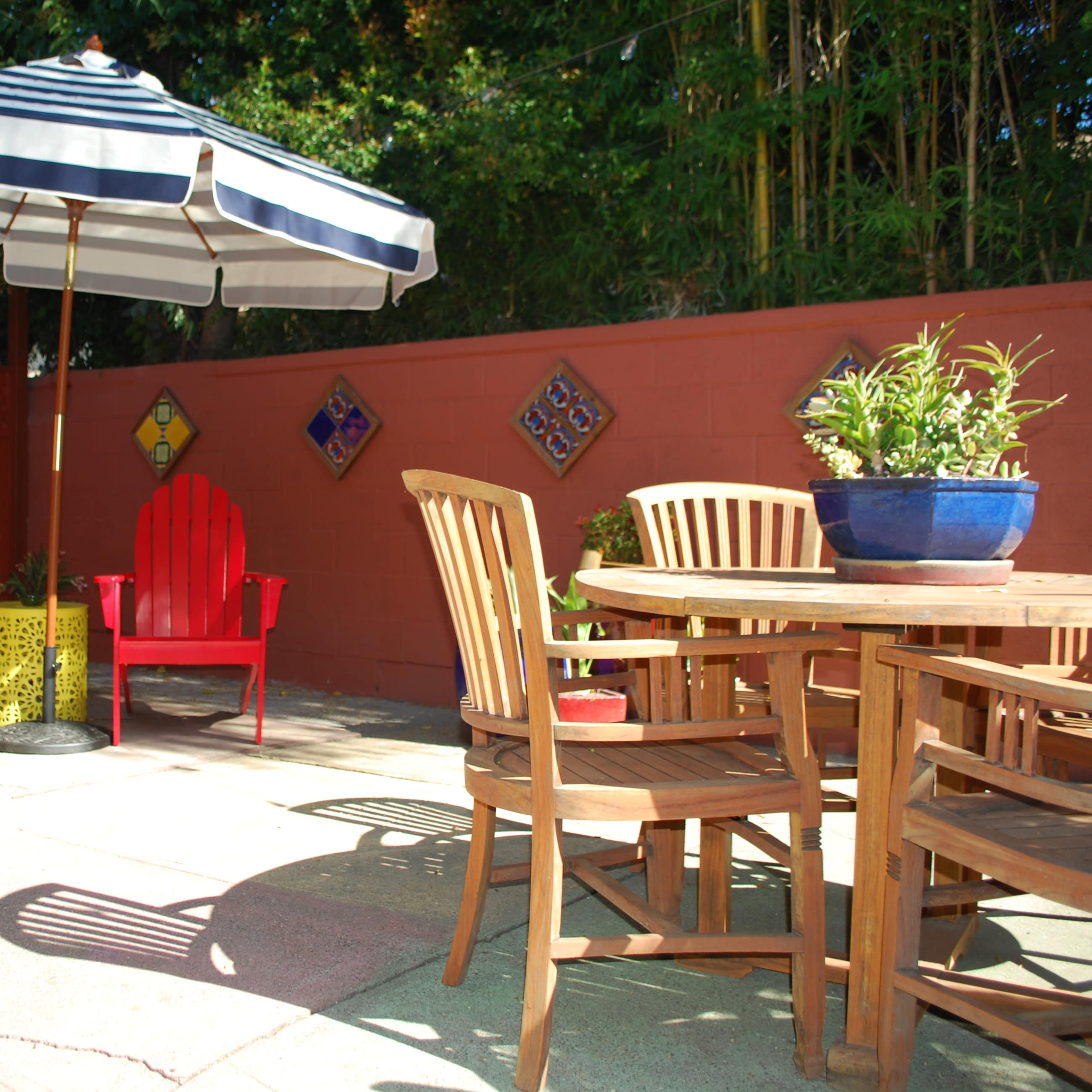 A small outdoor patio features a round wooden table surrounded by chairs, with two colorful adirondack chairs and a large striped umbrella in a corner.