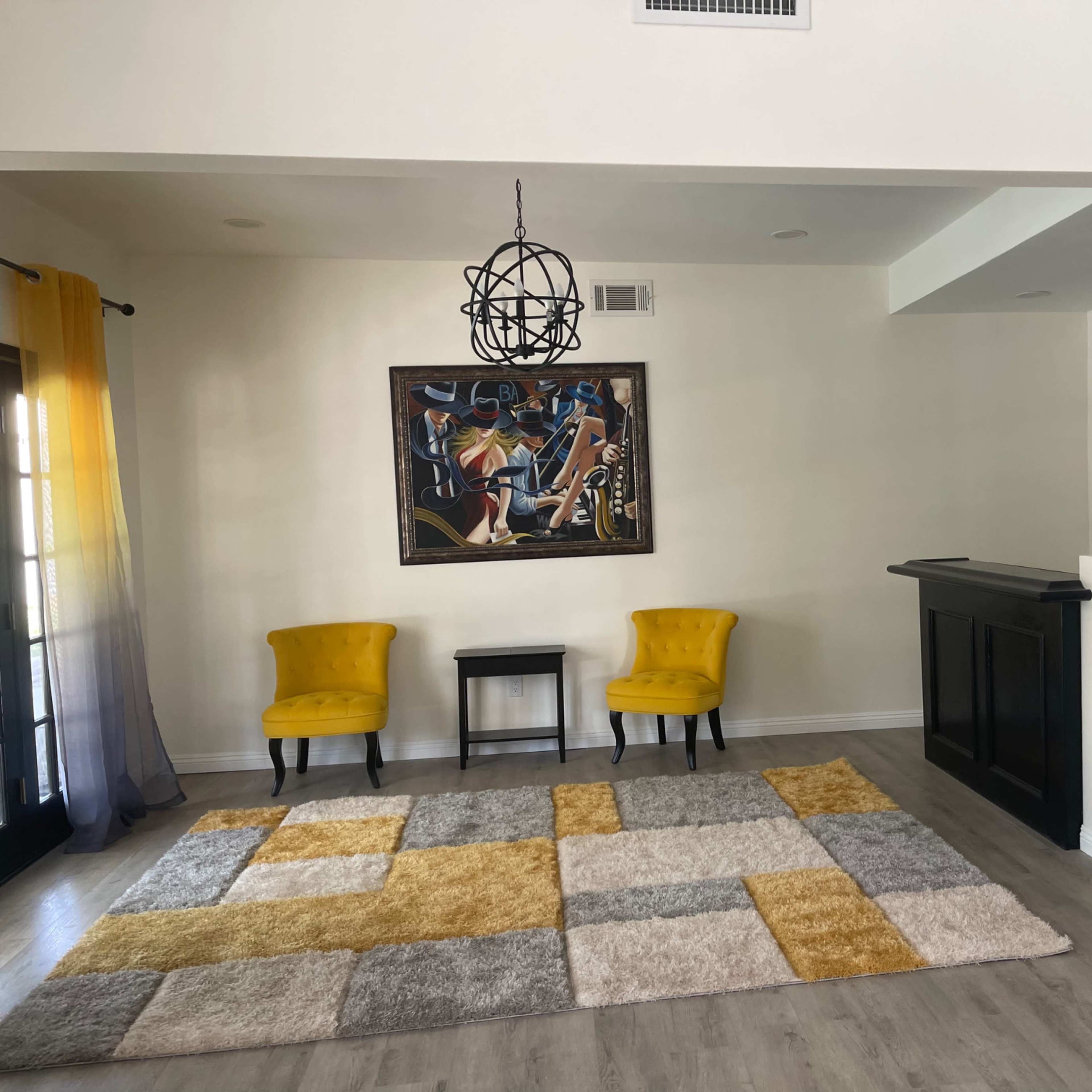 A bright living area featuring two yellow chairs, a small black table, a patterned rug, and a framed artwork on the wall.