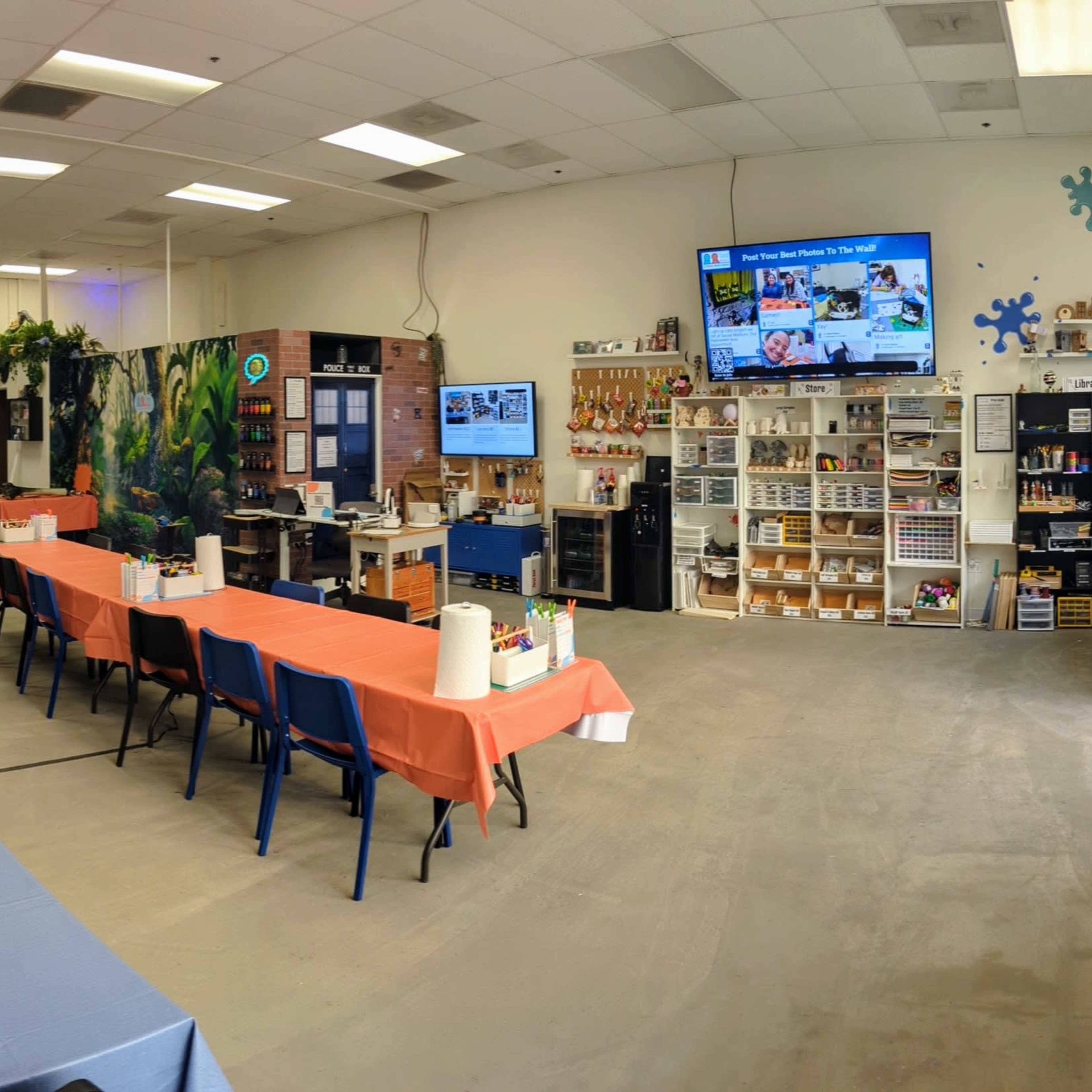 A spacious art studio with multiple tables set for painting activities, two large screens displaying content, and various art supplies organized on shelves.
