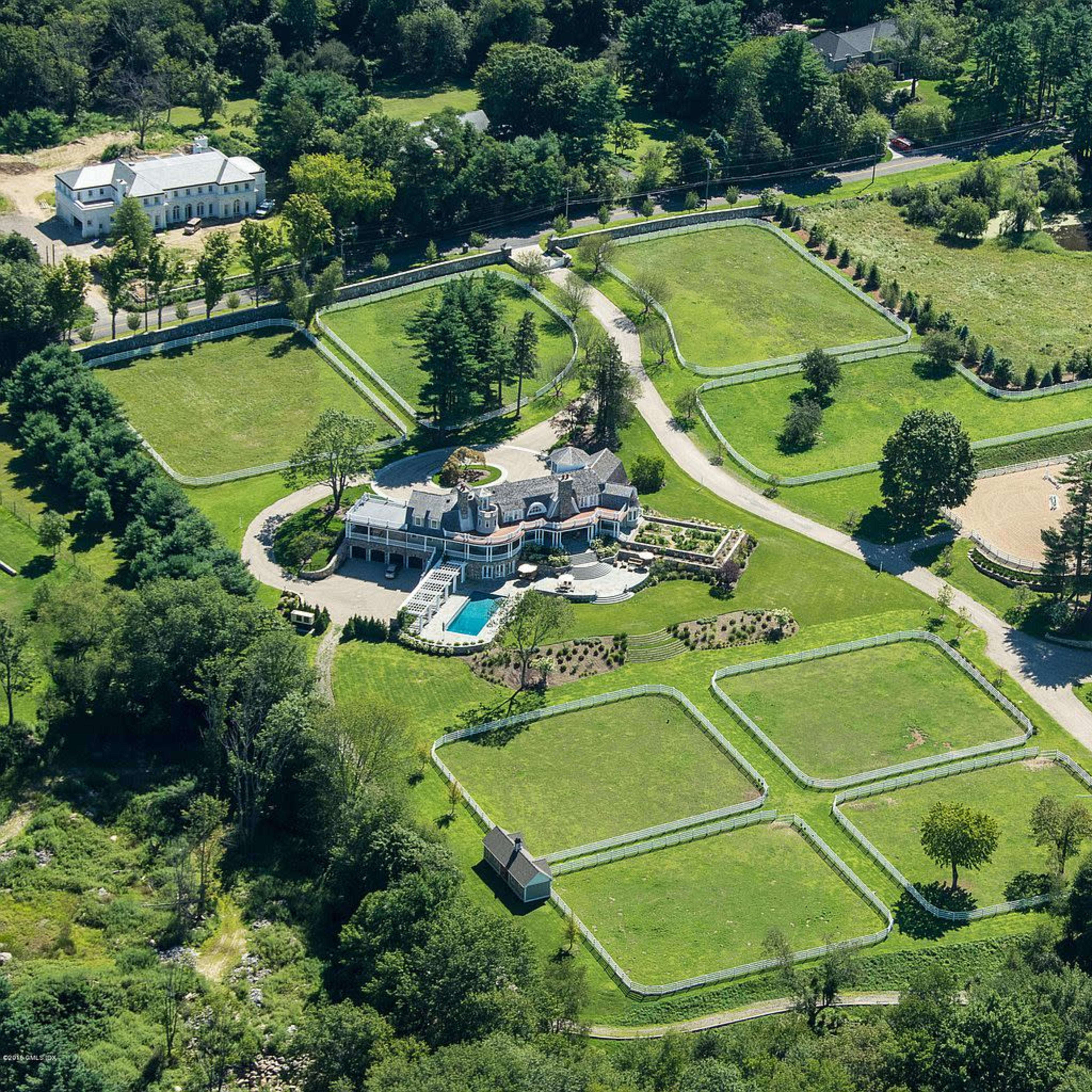 A large estate with manicured lawns, a swimming pool, and several buildings situated on expansive green grounds bordered by trees.