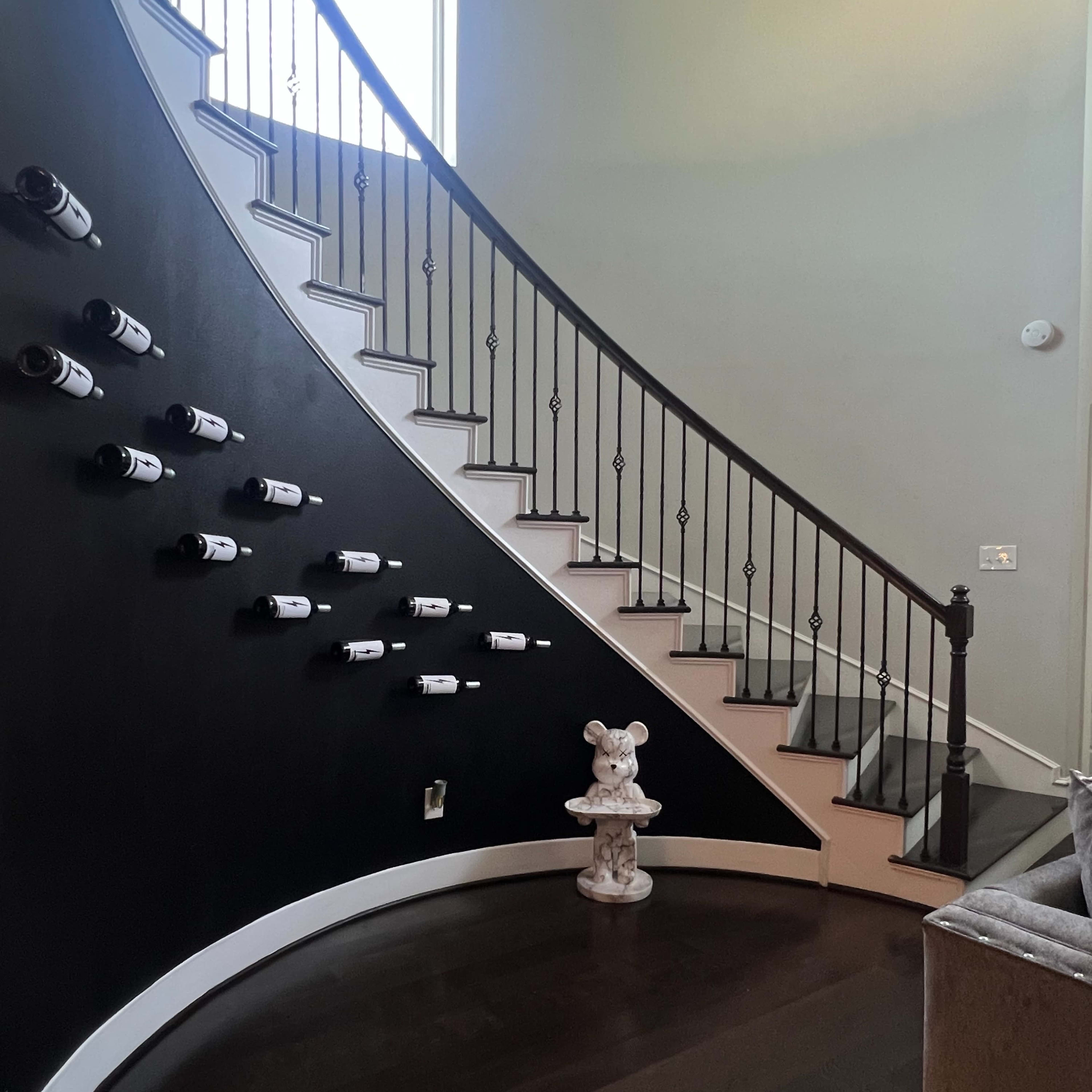 A curved staircase with a black wall displays wine bottles, while a decorative statue stands at the bottom.