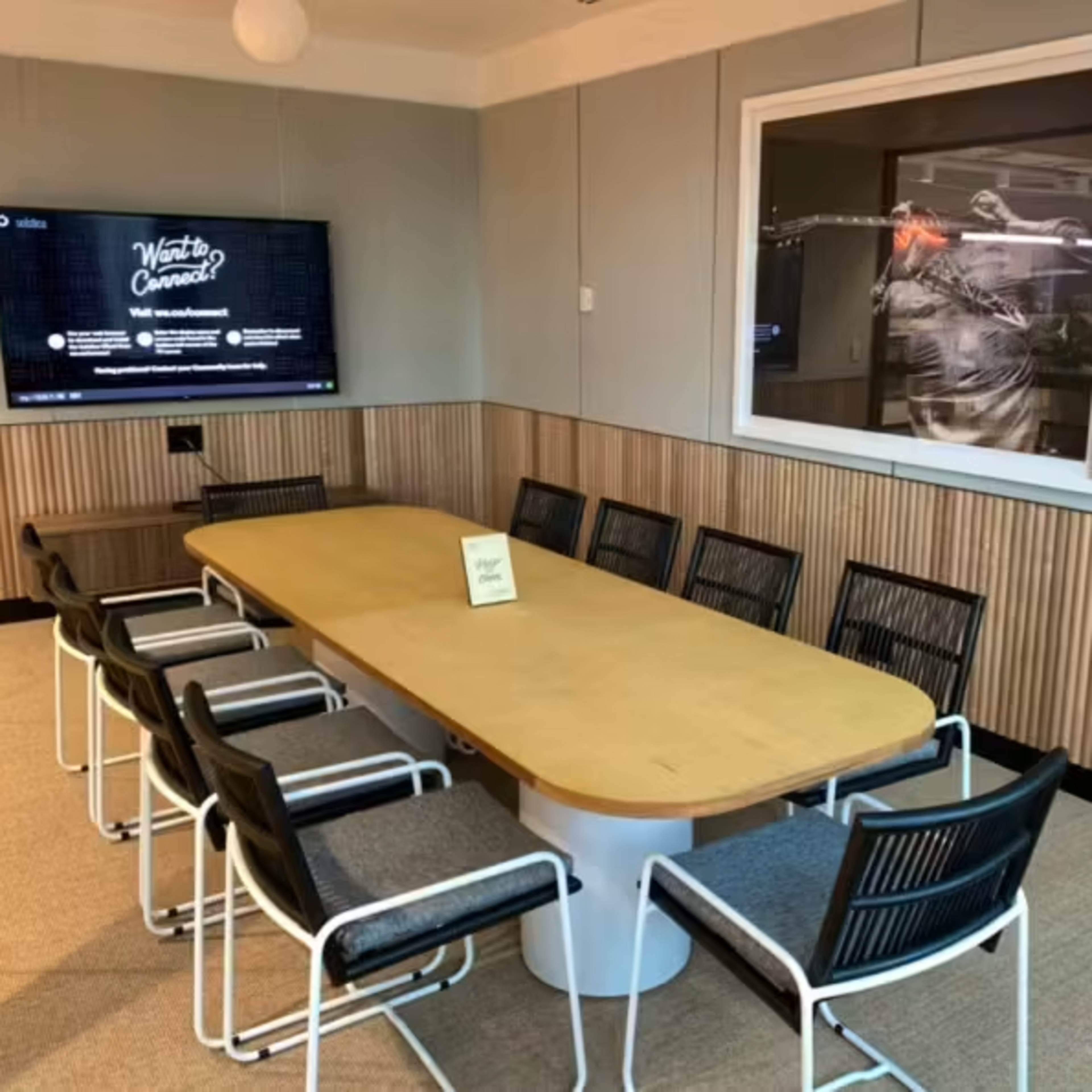 A large conference room features a long wooden table surrounded by several chairs, with a television mounted on the wall and a framed picture hanging nearby.