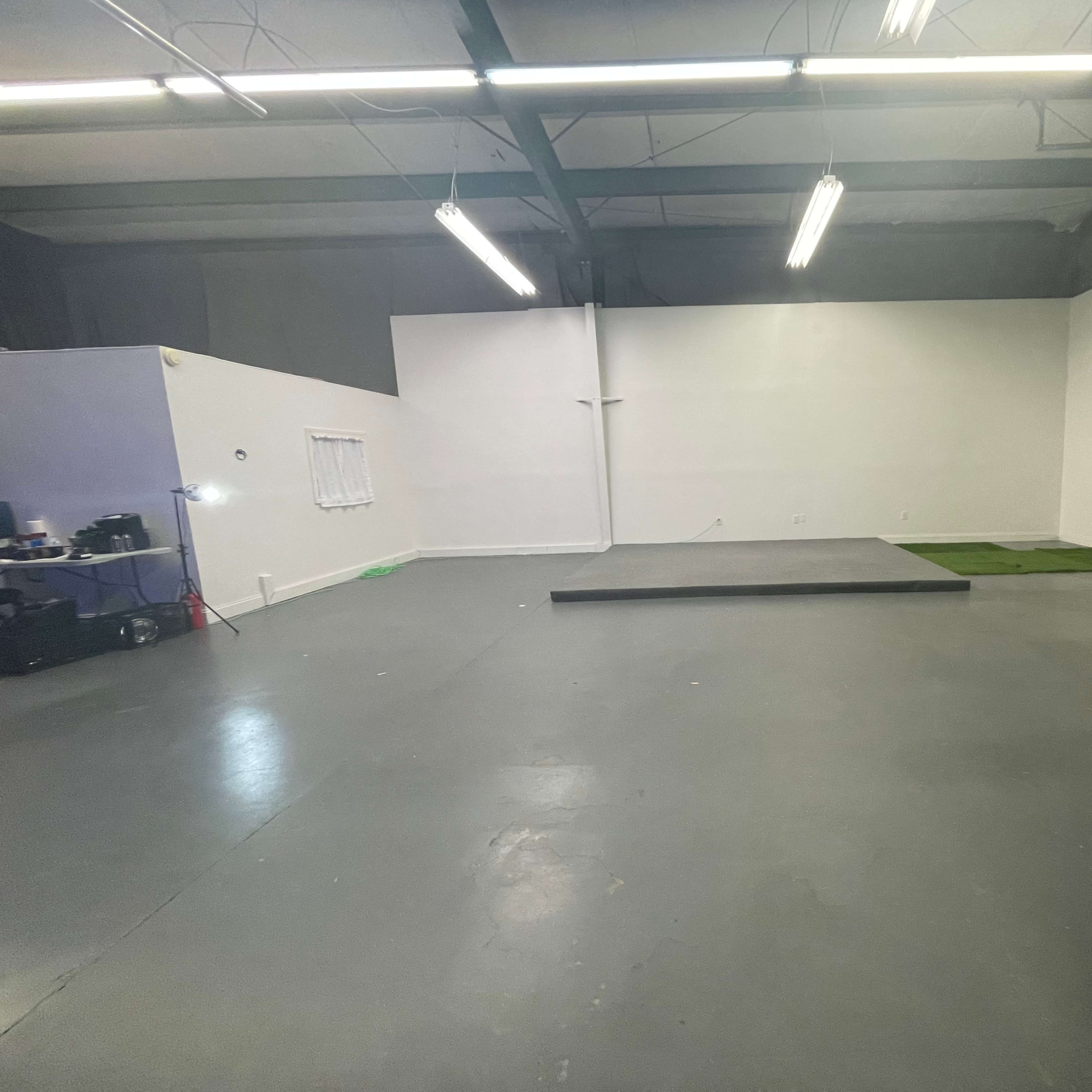 An empty room with a gray floor, a raised platform at the center, and white walls, illuminated by overhead fluorescent lights.