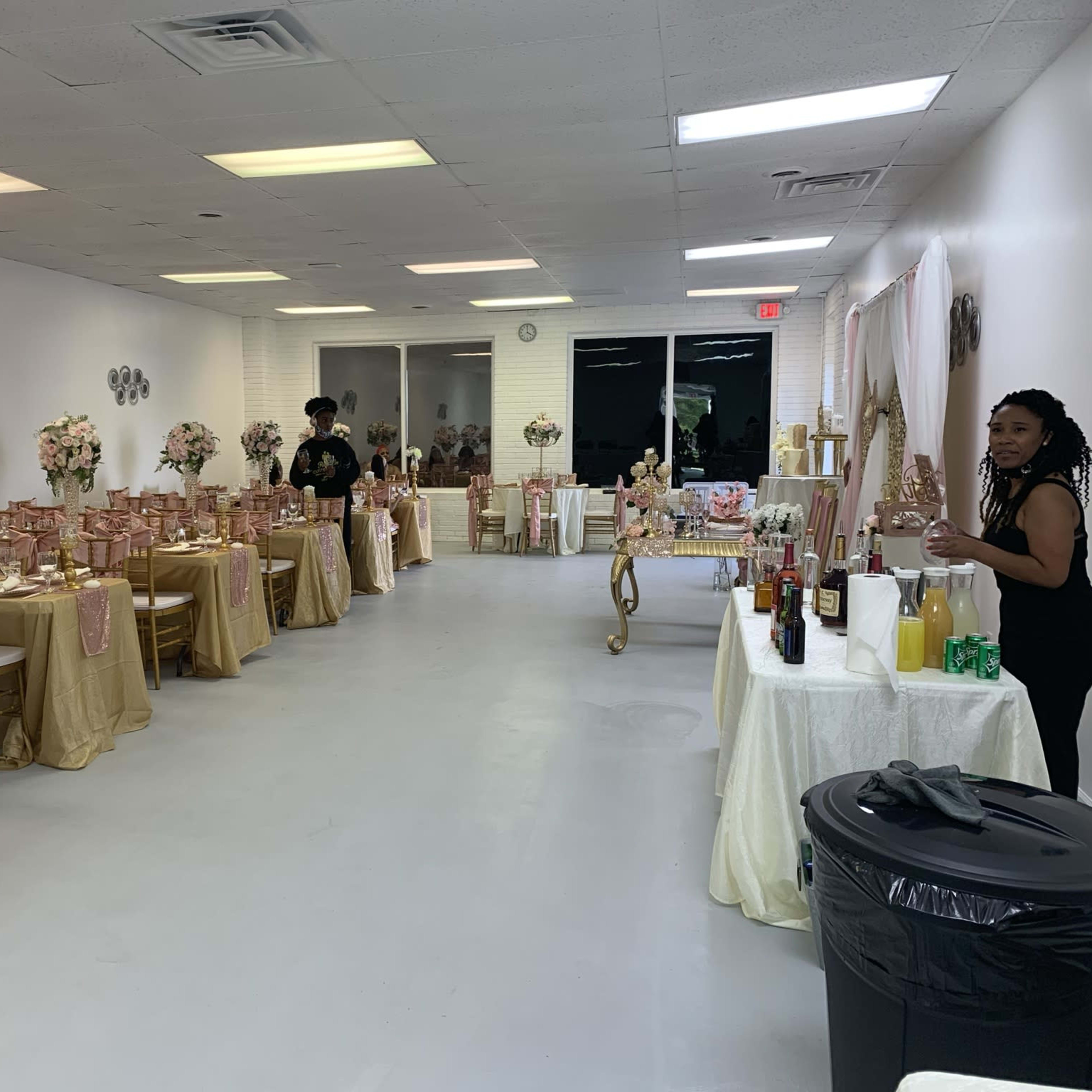 A decorated event space arranged with tables draped in gold and pink tablecloths, floral centerpieces, and a beverage station along one side.