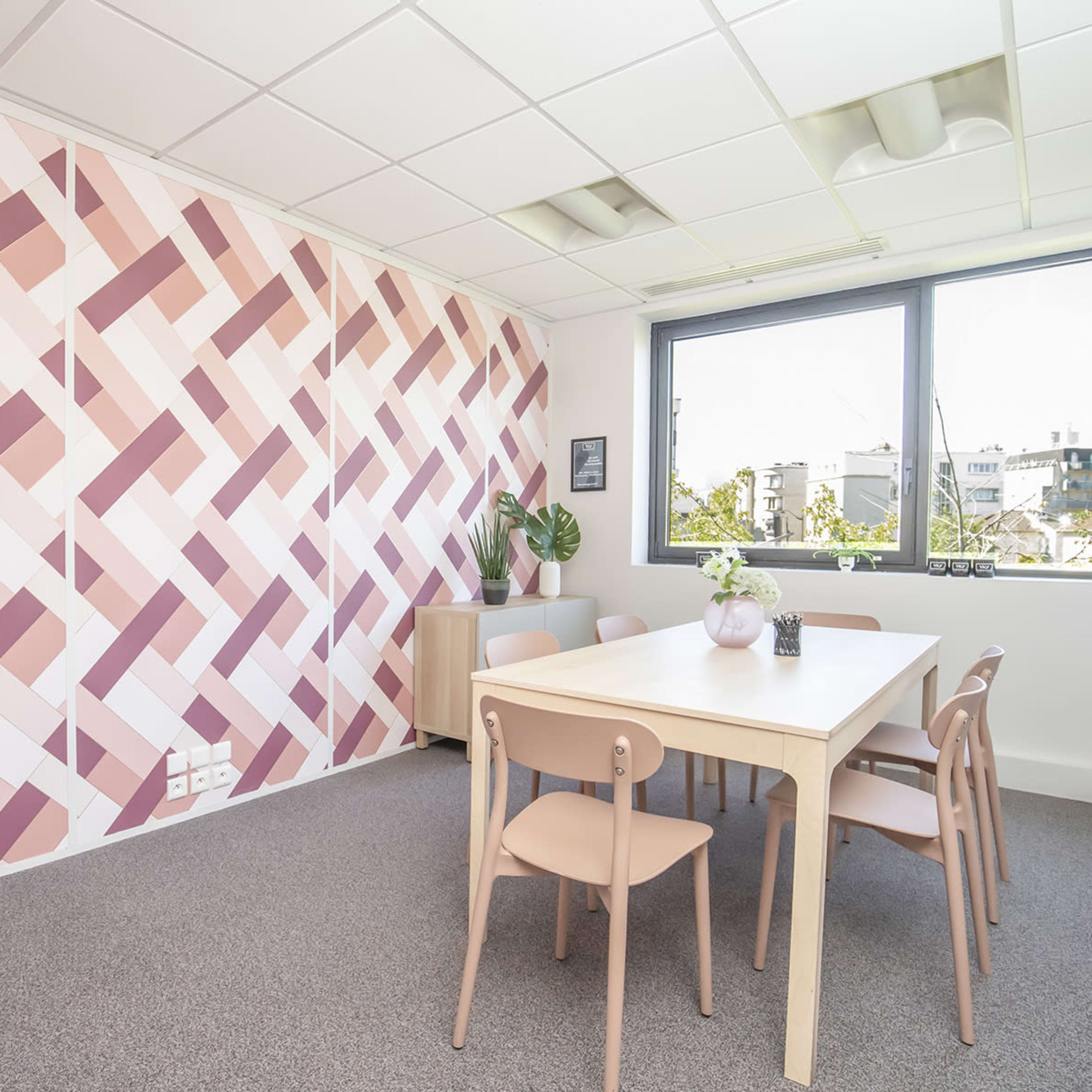 A modern office space featuring a table surrounded by chairs, a potted plant, and a patterned pink and white wall.