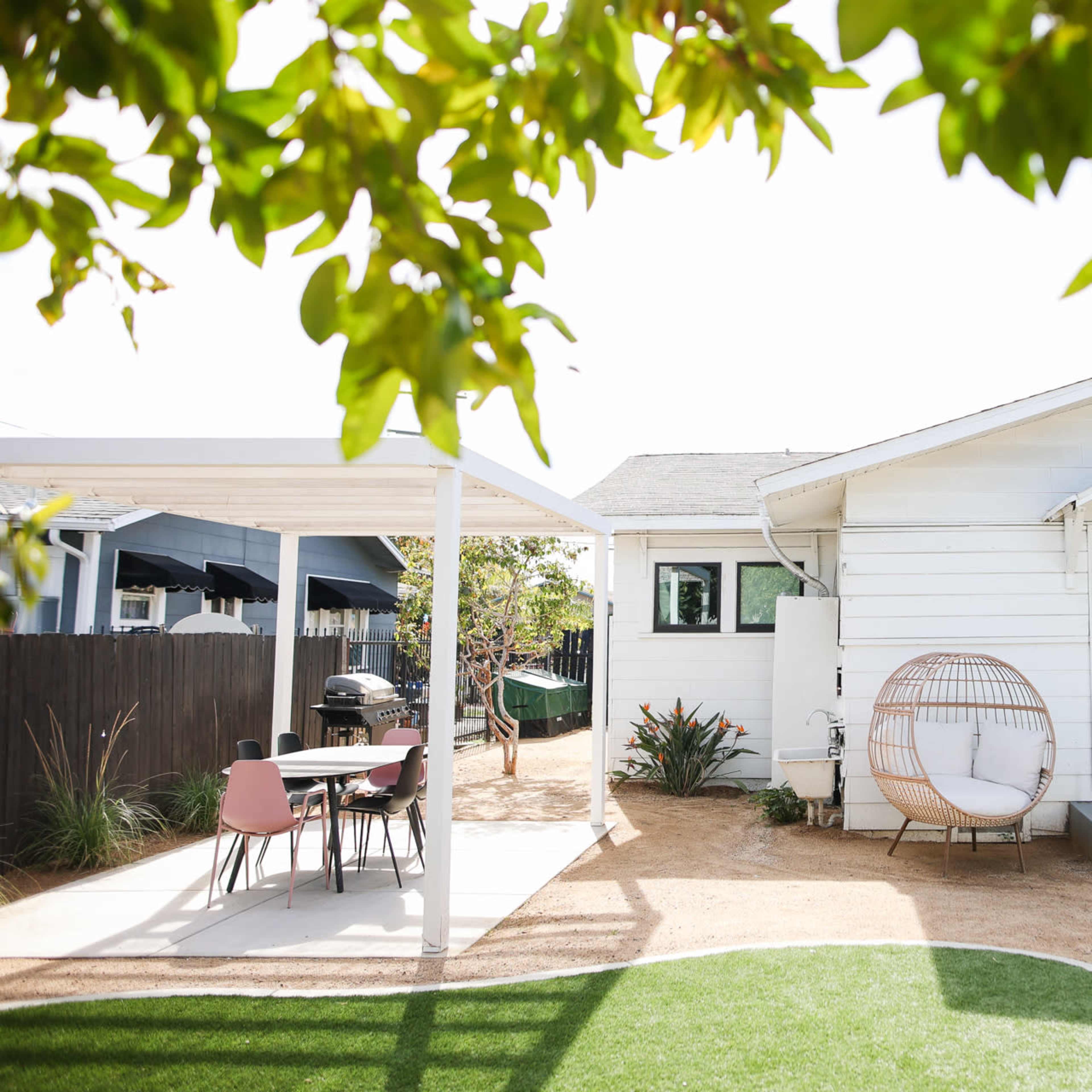 A white house with a front yard featuring a circular seating area, a dining table under a pergola, and a lemon tree with ripe lemons.
