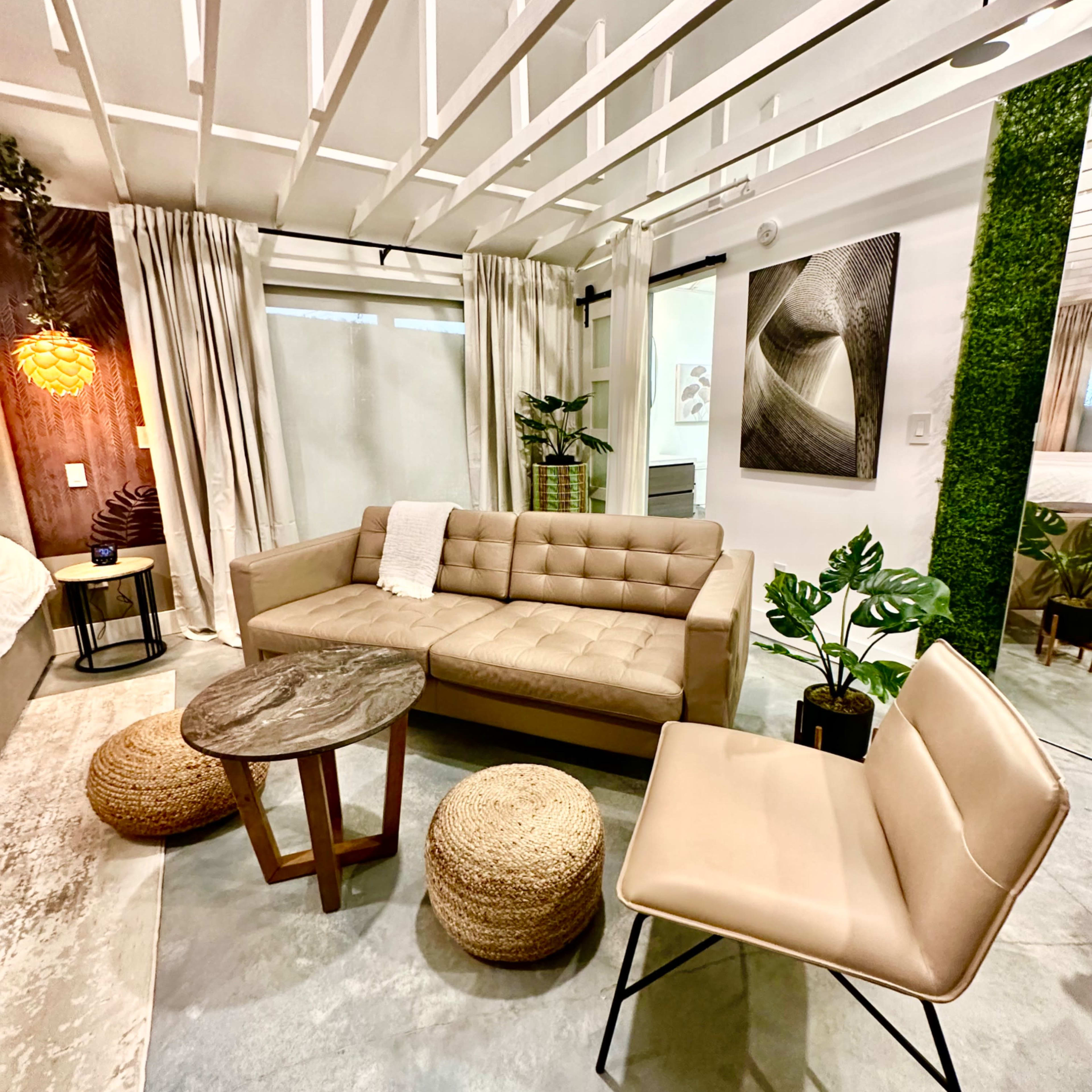 A modern living space featuring a beige sofa, a round wooden coffee table, two textured ottomans, and a sleek chair, surrounded by green plants and stylish decor.