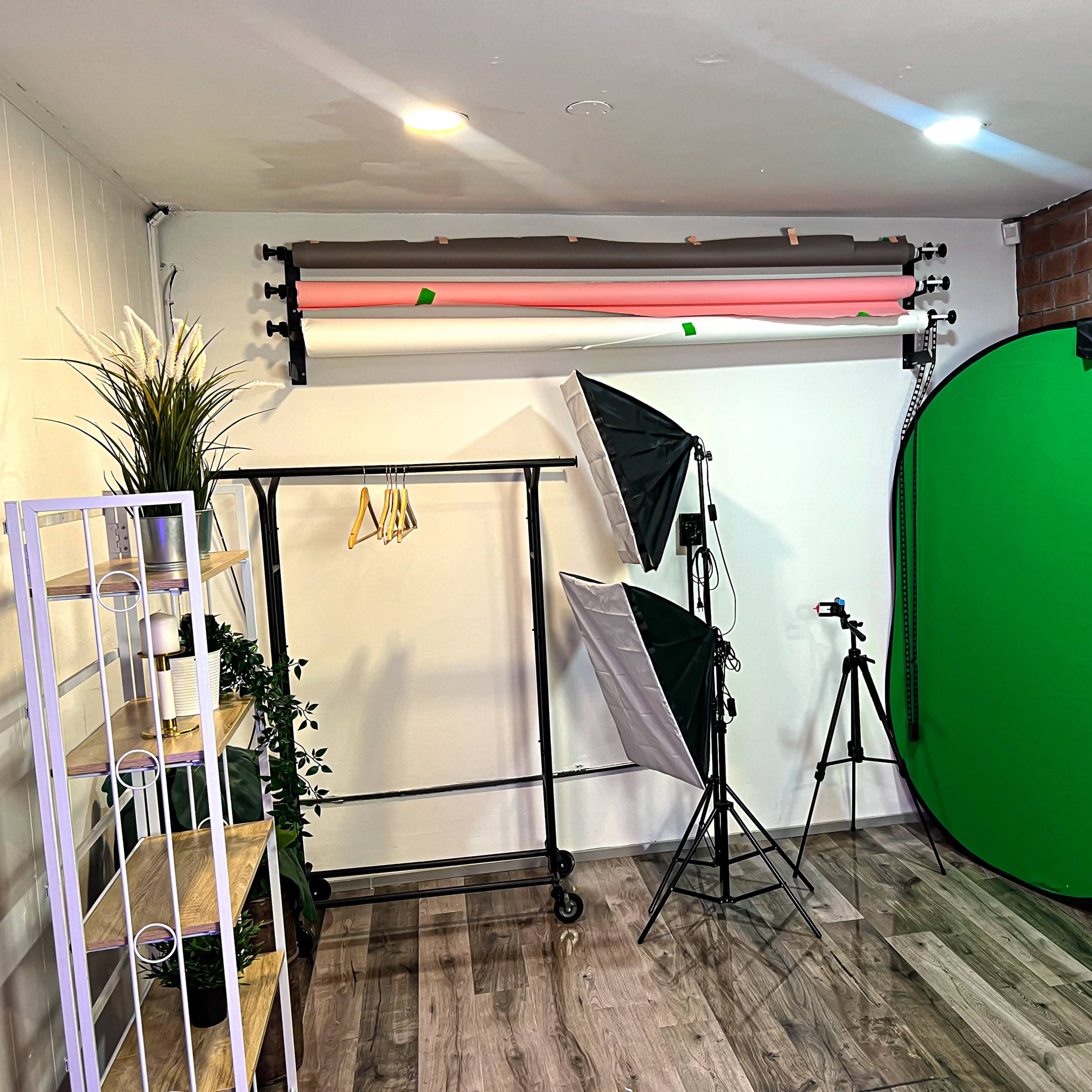 A photography studio setup with a green backdrop, lighting equipment, a clothing rack, and a shelf against a textured wall.