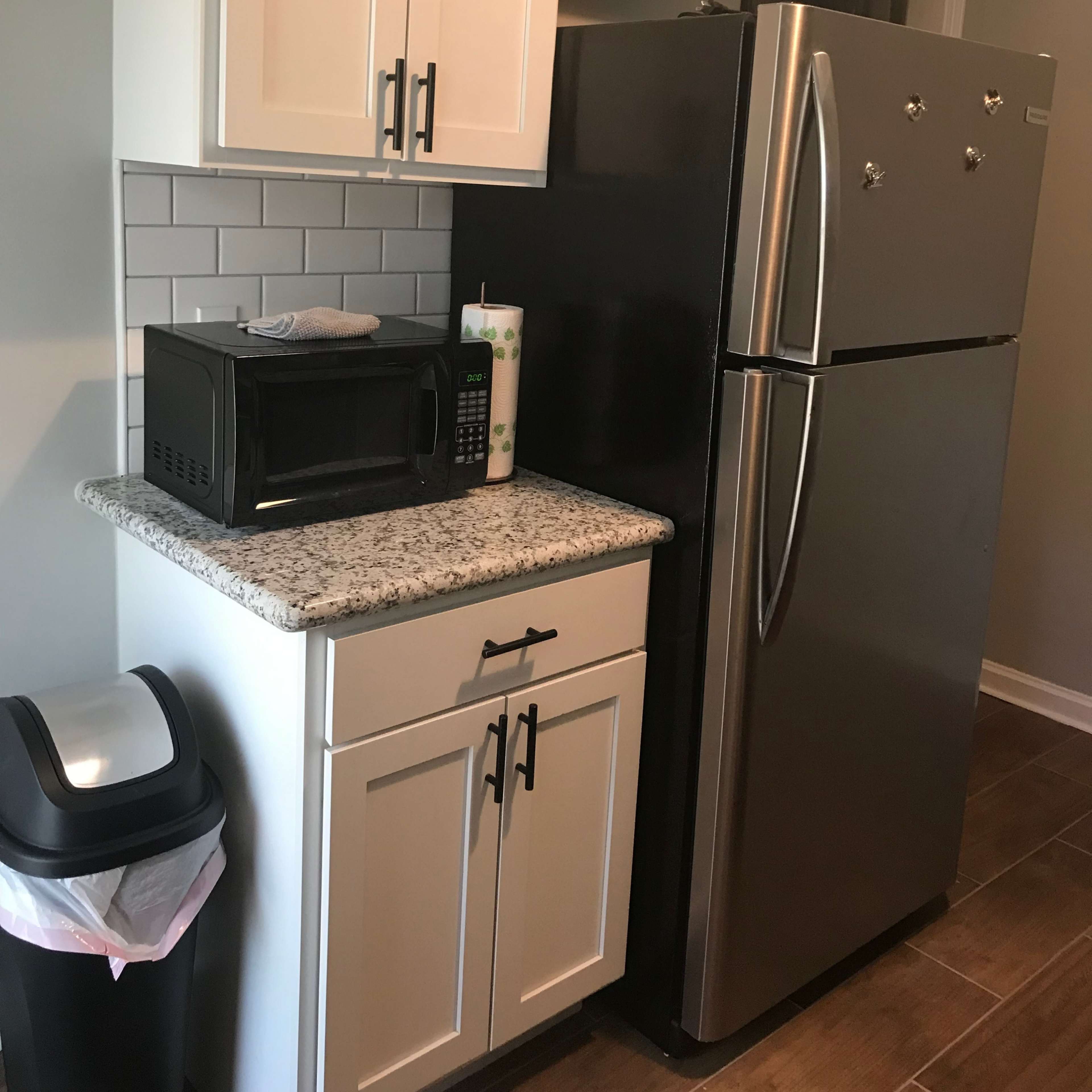 A kitchen corner featuring a granite countertop, a black microwave, a stainless steel refrigerator, and a trash can.