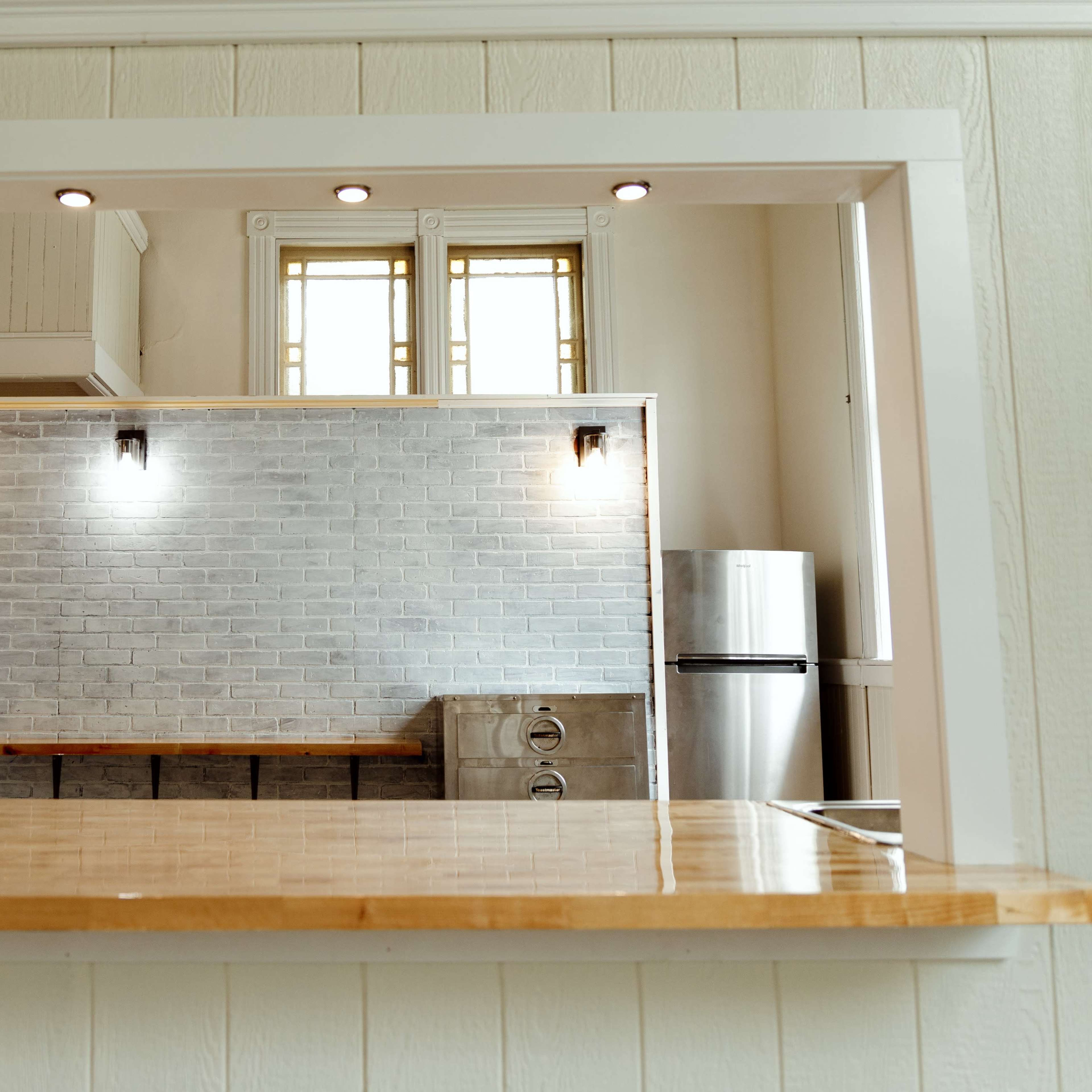 A kitchen with a wooden countertop and a grey brick wall, featuring a stainless steel refrigerator and overhead lighting.