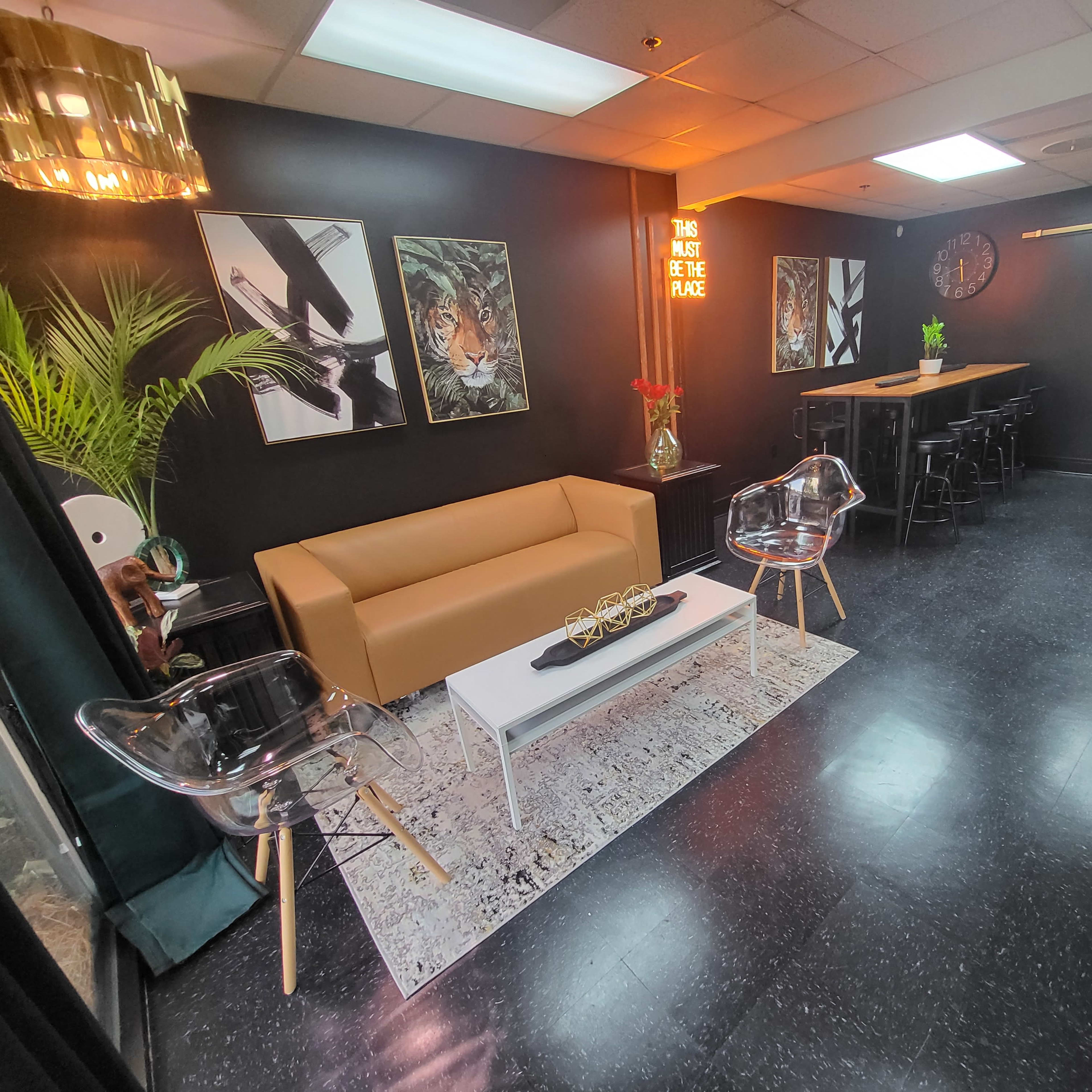 A modern lounge area with a brown leather sofa, transparent chairs, a white coffee table, and a black bar with tall stools against a dark wall, decorated with abstract artwork and greenery.