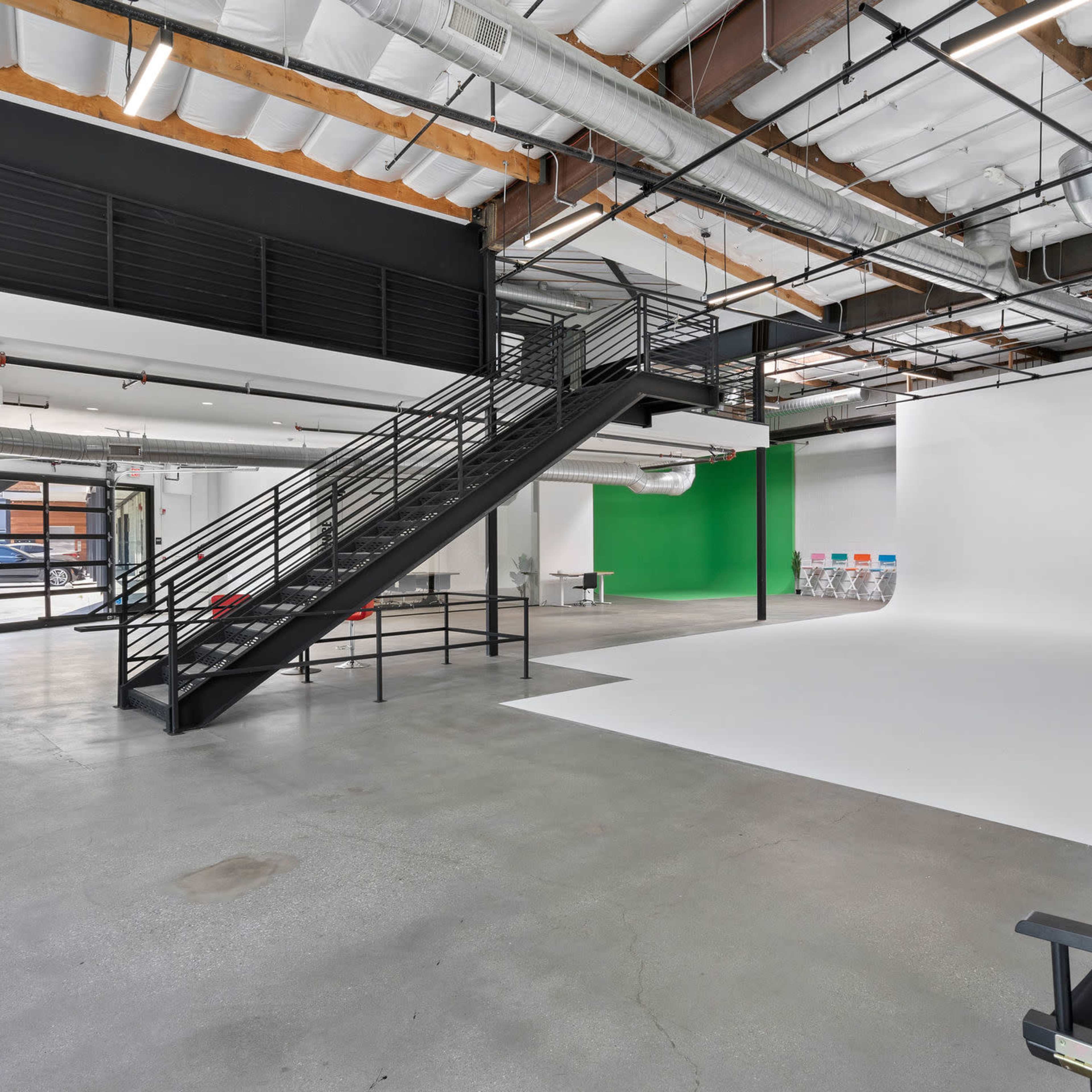 A spacious studio with a large white backdrop, a green wall, a black staircase, and several chairs arranged in front of the backdrop.