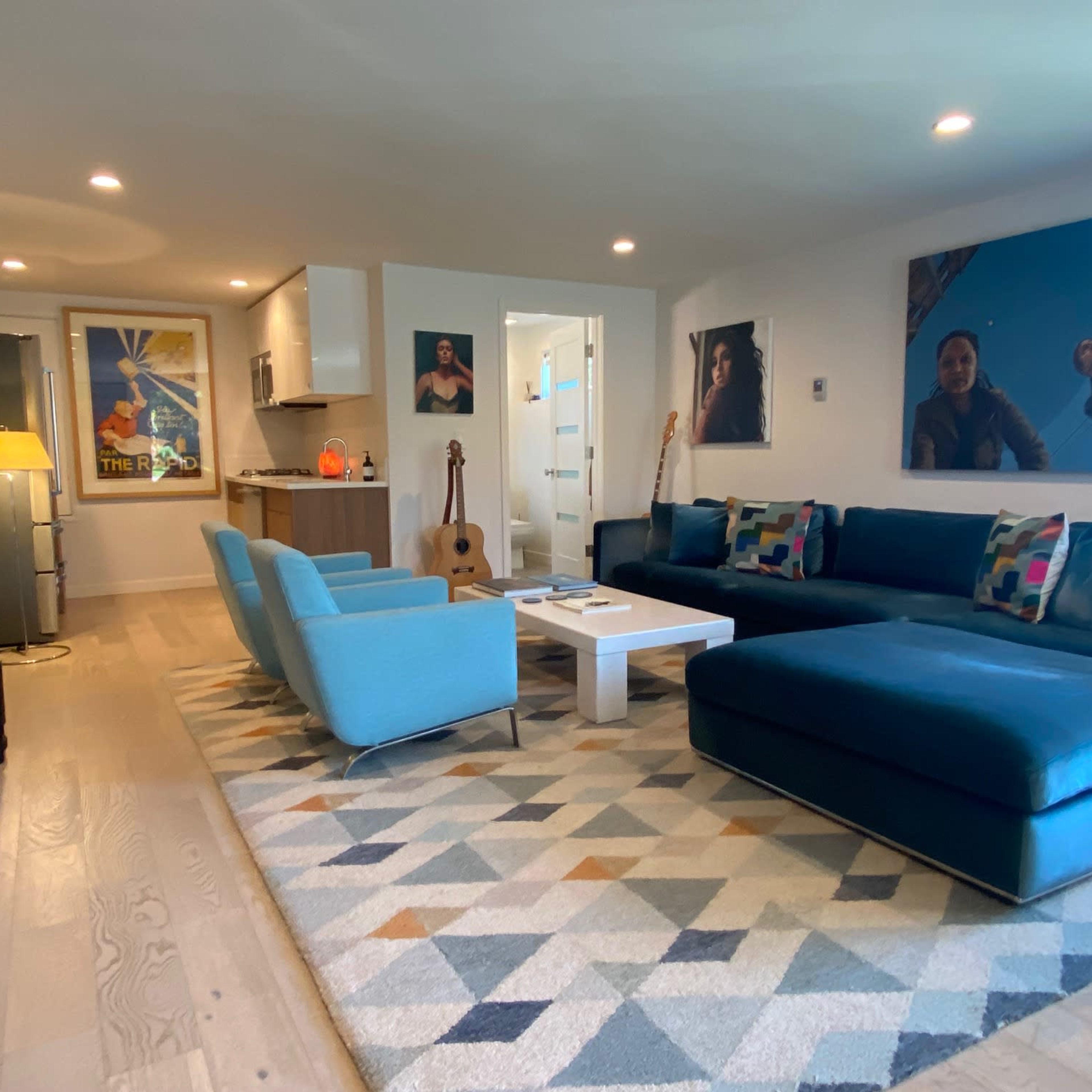 The living room features a blue sectional sofa, a coffee table, a patterned rug, and various artworks on the walls, with a bookshelf and a guitar nearby.