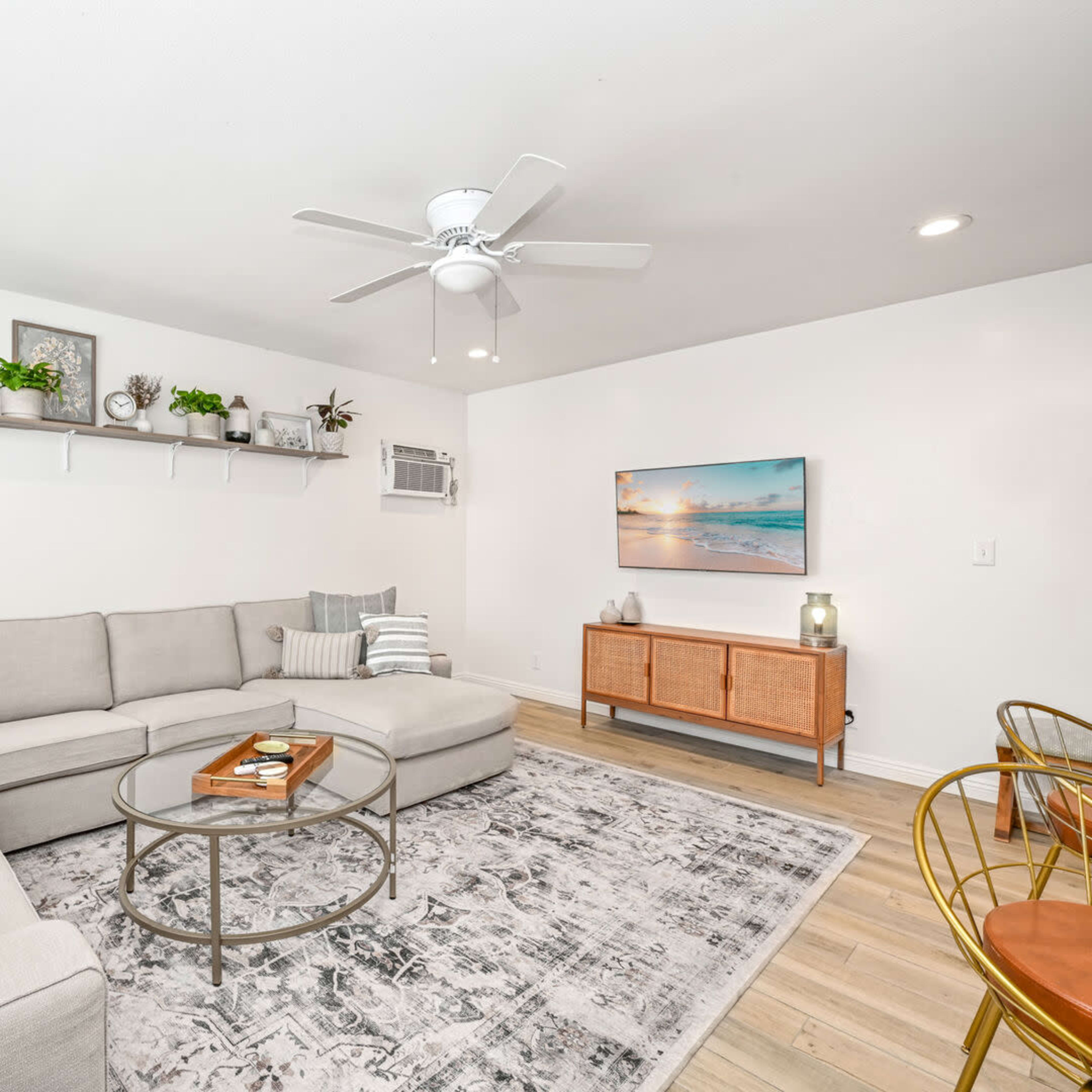 A modern living room features a sectional sofa, coffee table, and a TV mounted on the wall, with plants on a shelf and wooden furniture.