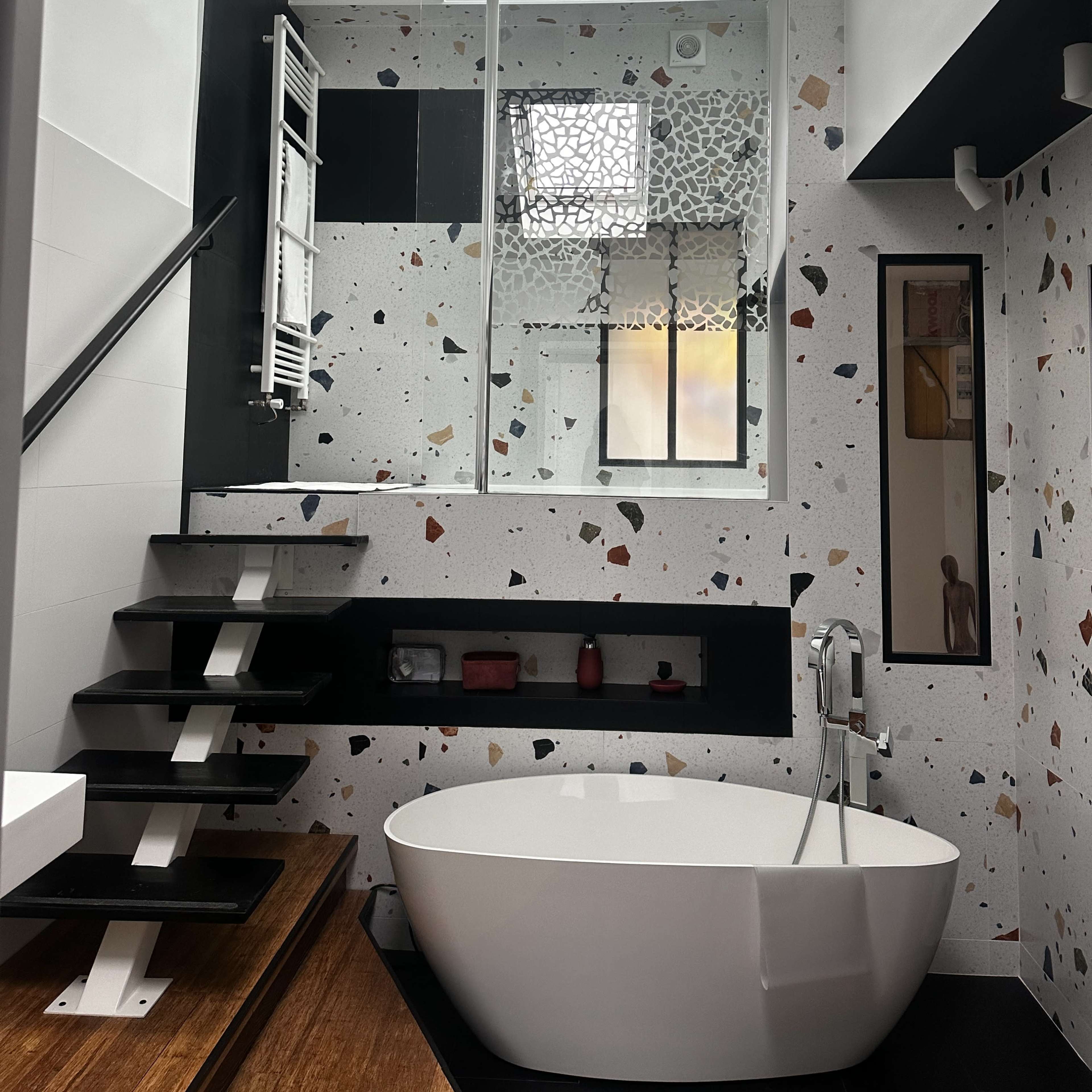 The bathroom features a freestanding bathtub, a glass-enclosed shower, and a staircase, all set against a patterned terrazzo wall and large skylight.