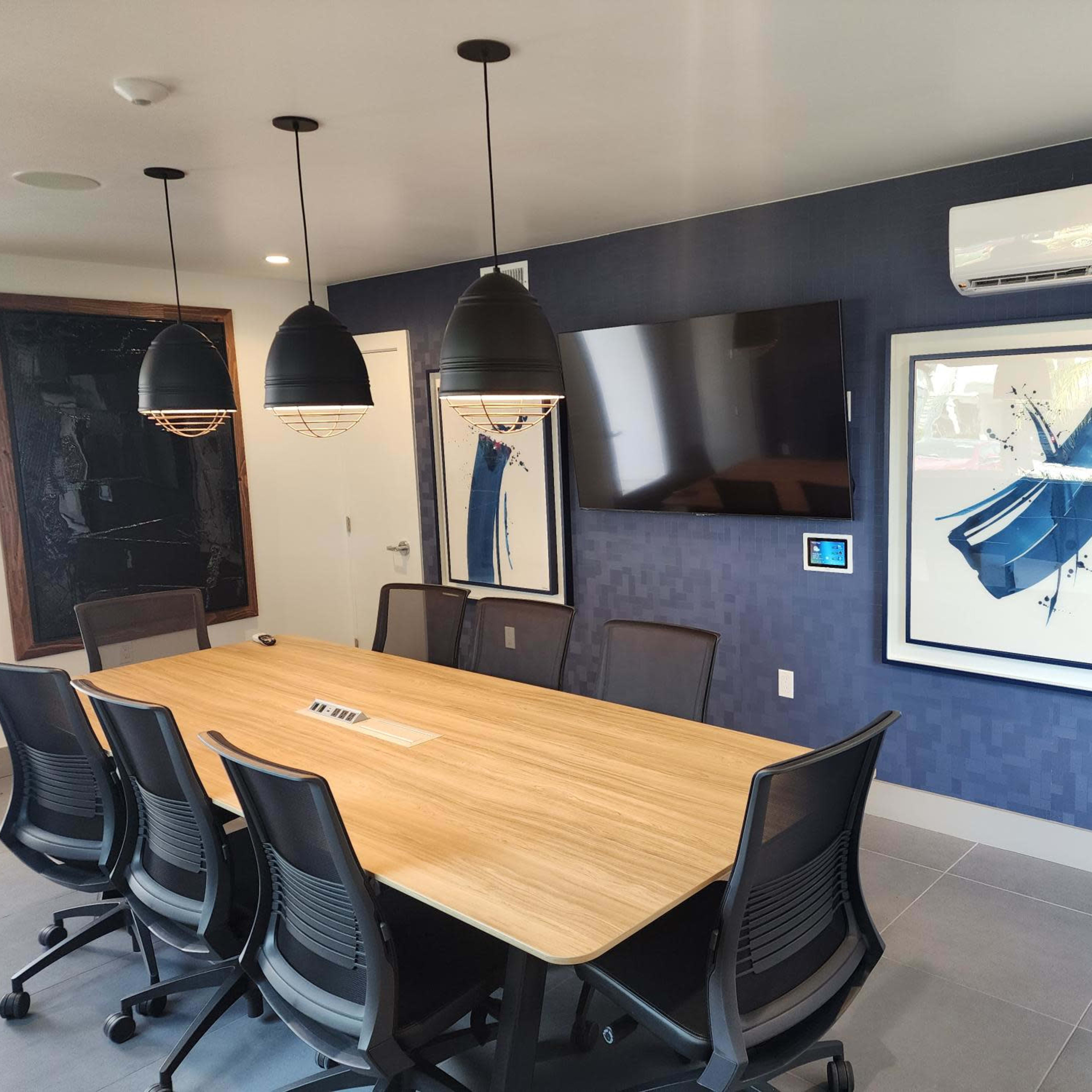 A modern conference room features a large wooden table surrounded by six black chairs, with two framed artworks and a wall-mounted television.