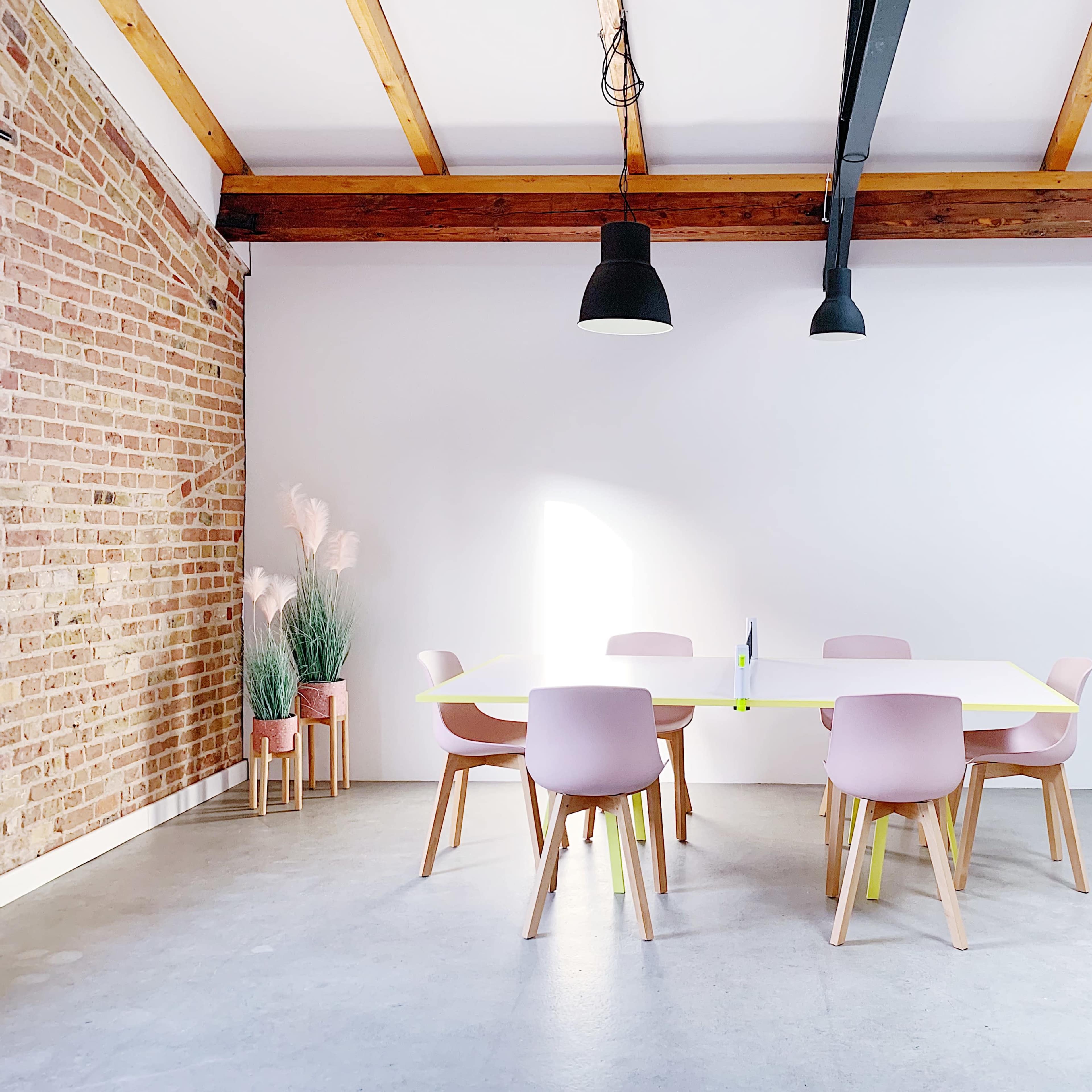 A spacious room features a large, white table surrounded by pastel pink chairs, with a brick wall and potted plants in the background.