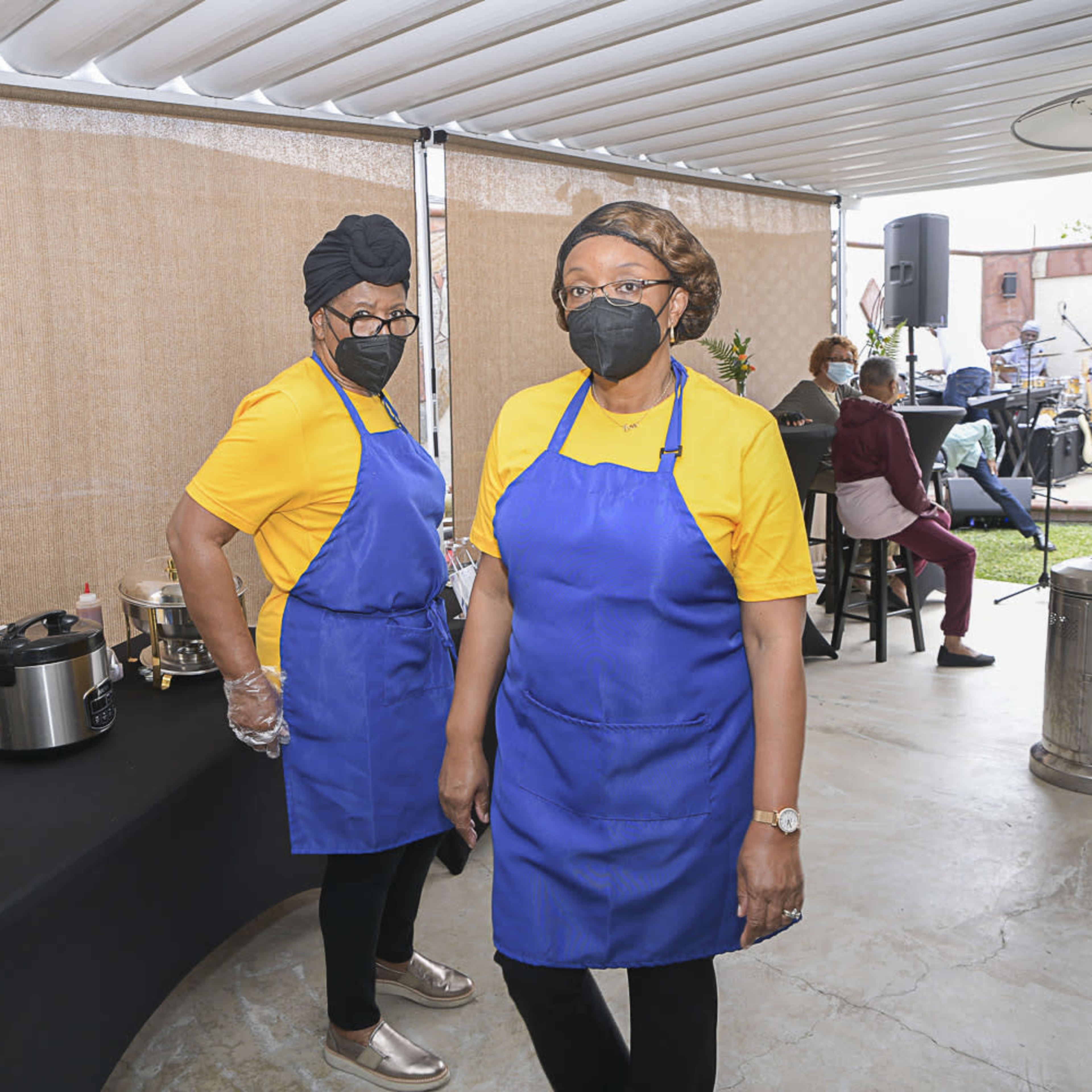 Two women wearing yellow shirts and blue aprons stand behind a table with food containers in an outdoor setting.