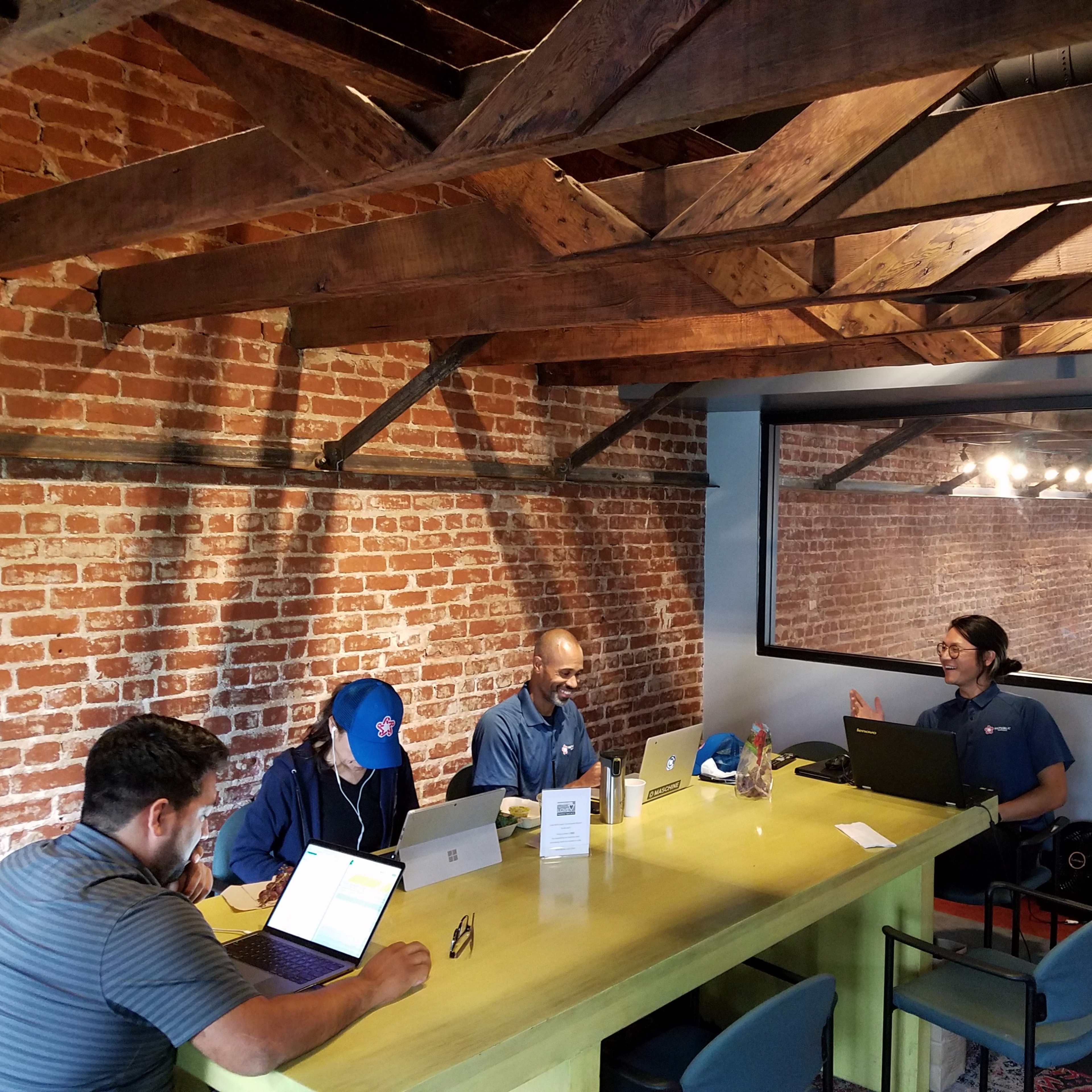A group of four people are working on laptops at a large table in a room with exposed brick walls and wooden beams.