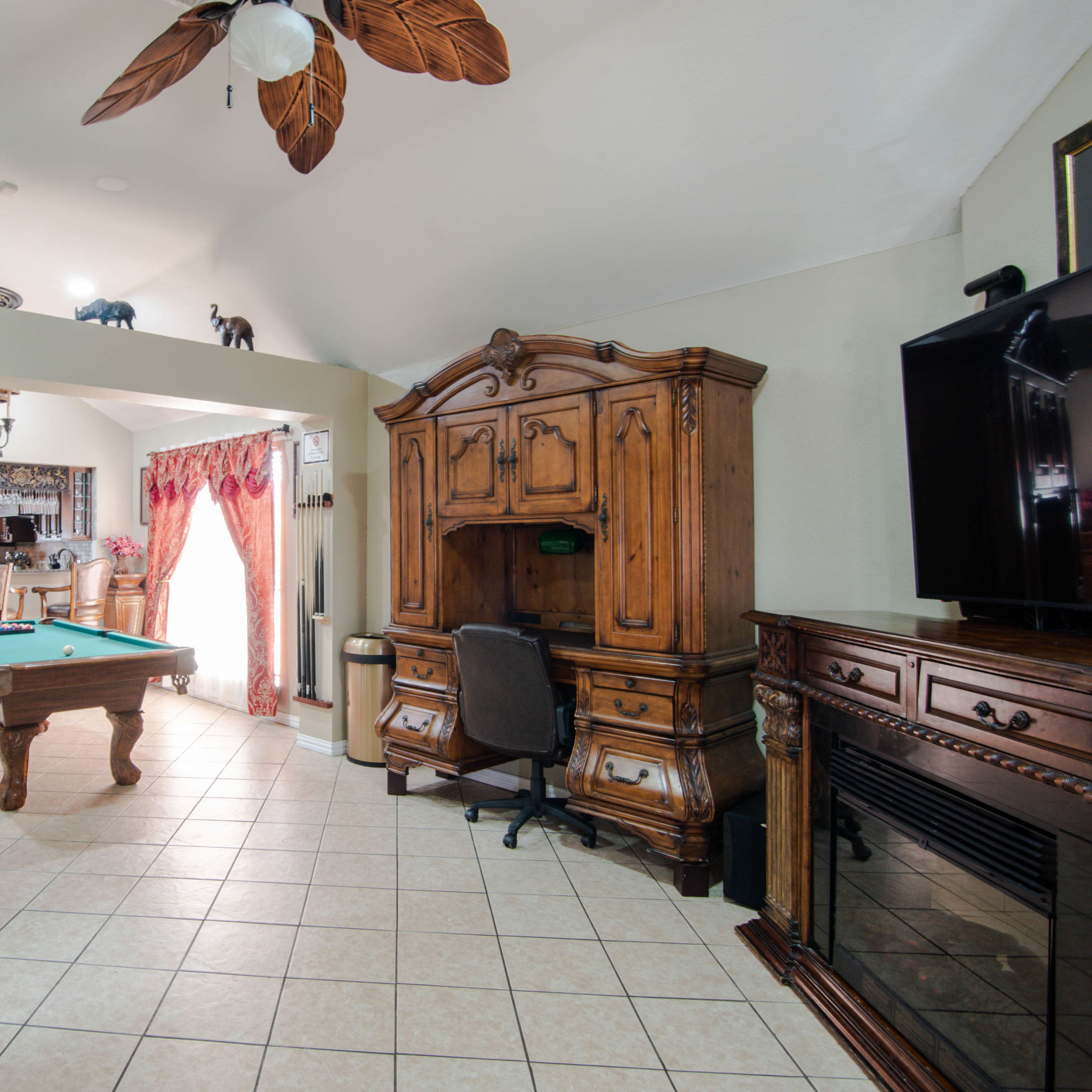 A spacious living room featuring a pool table, a wooden desk, and a large entertainment center, with decorative plants and warm lighting.
