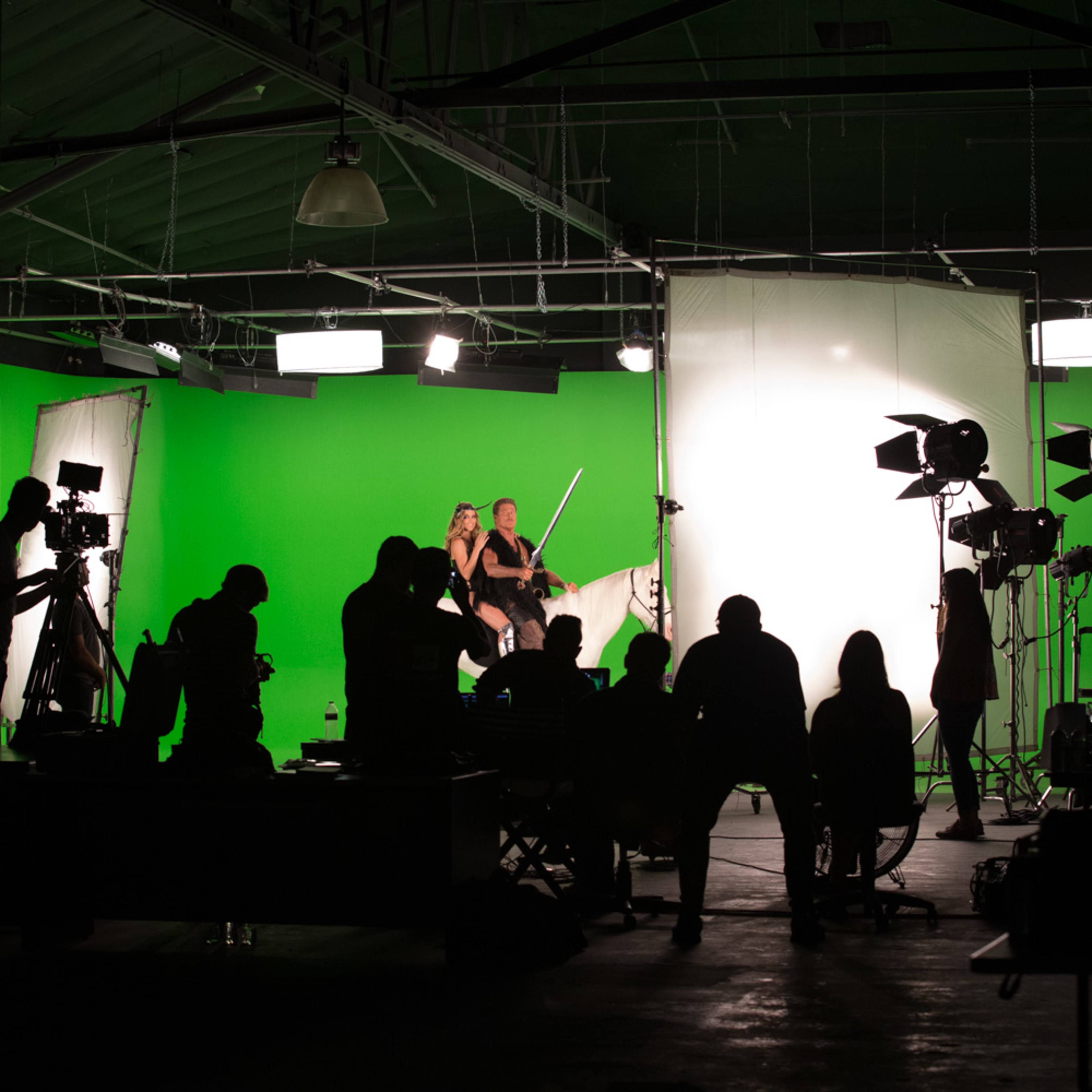 A film crew works in a studio with a green backdrop while silhouettes of people sit in the foreground.