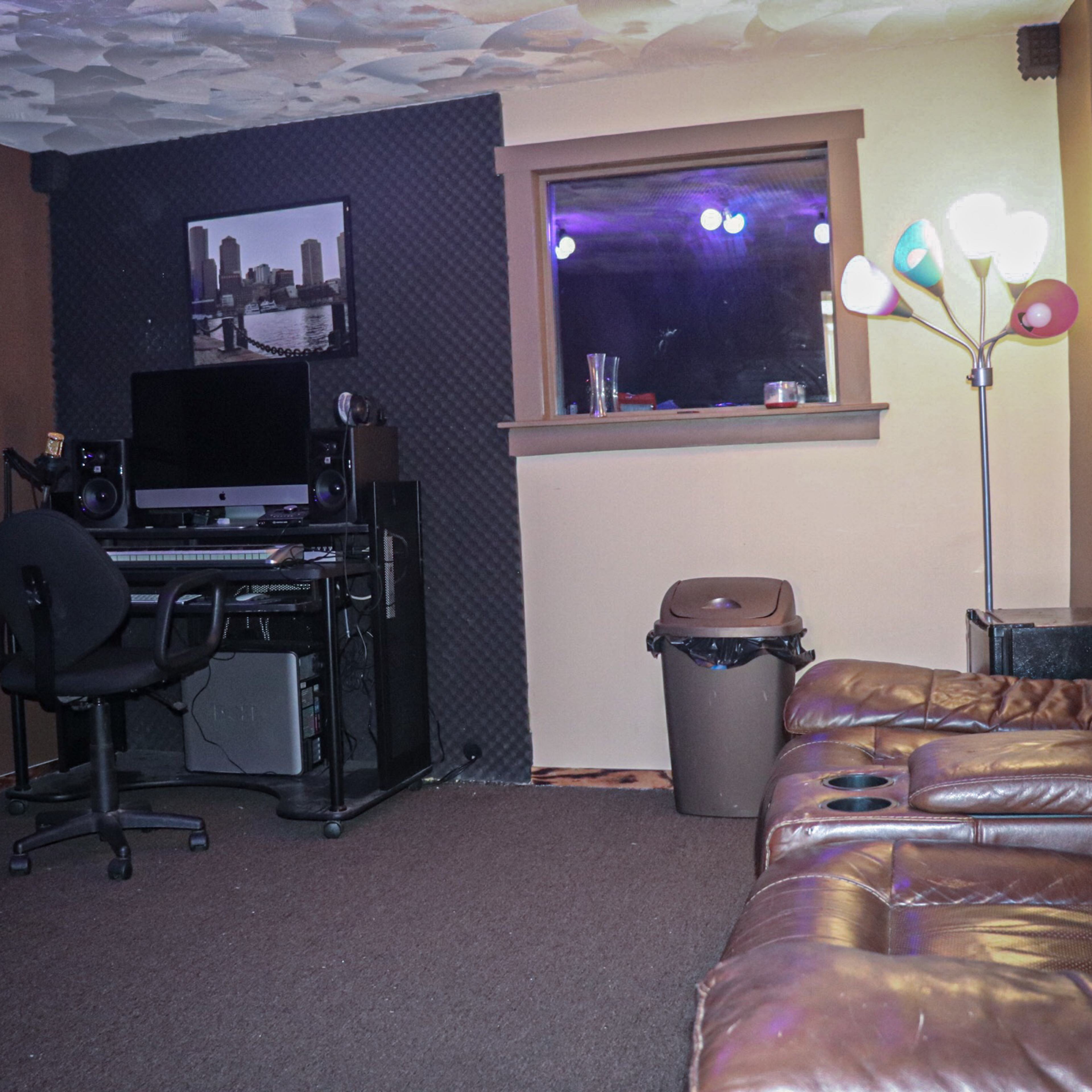 A corner of a room features a desk with a computer setup beside a window, brown leather recliners, and a trash can.