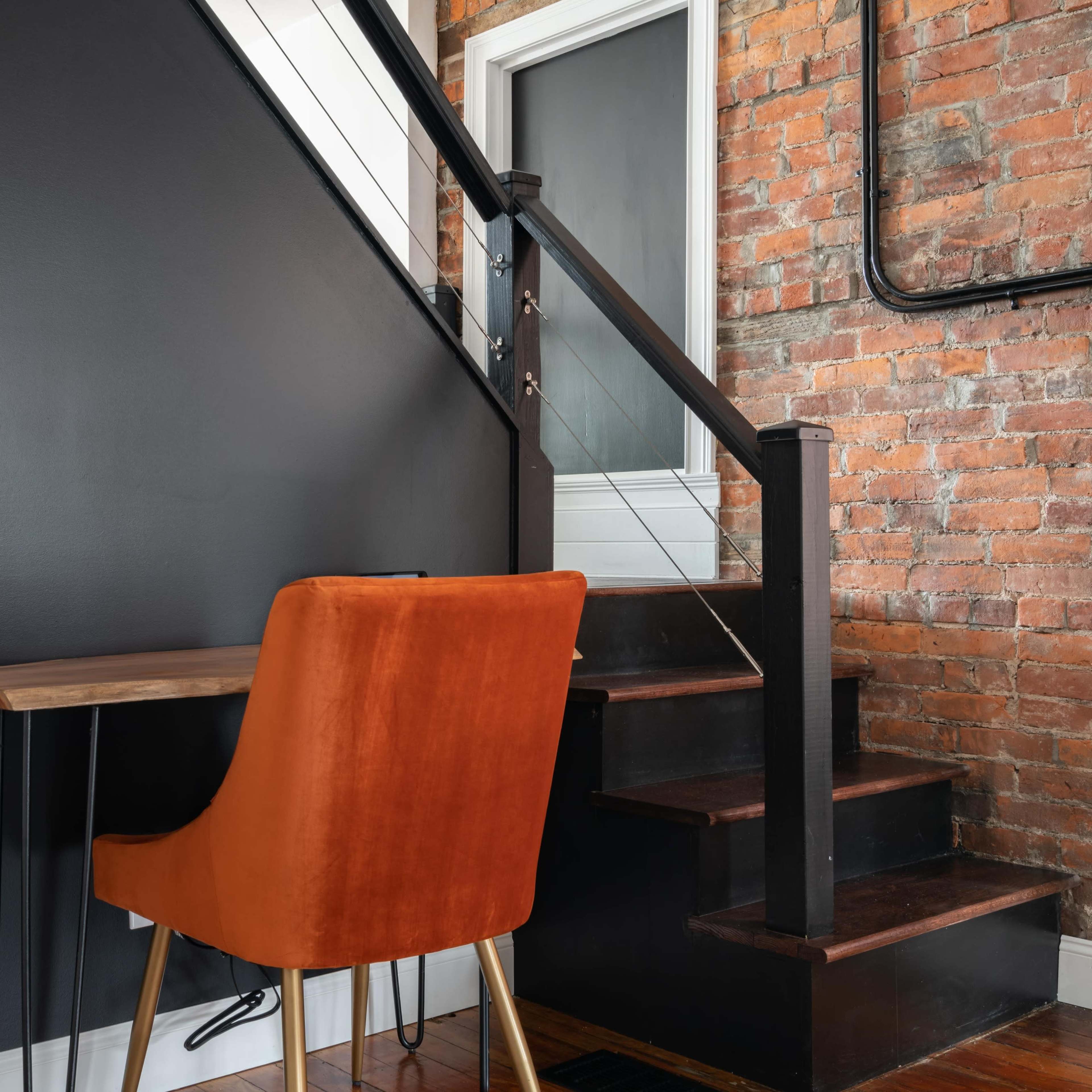 A small workspace with an orange chair and wooden table positioned against a black wall, next to a staircase with exposed brick.