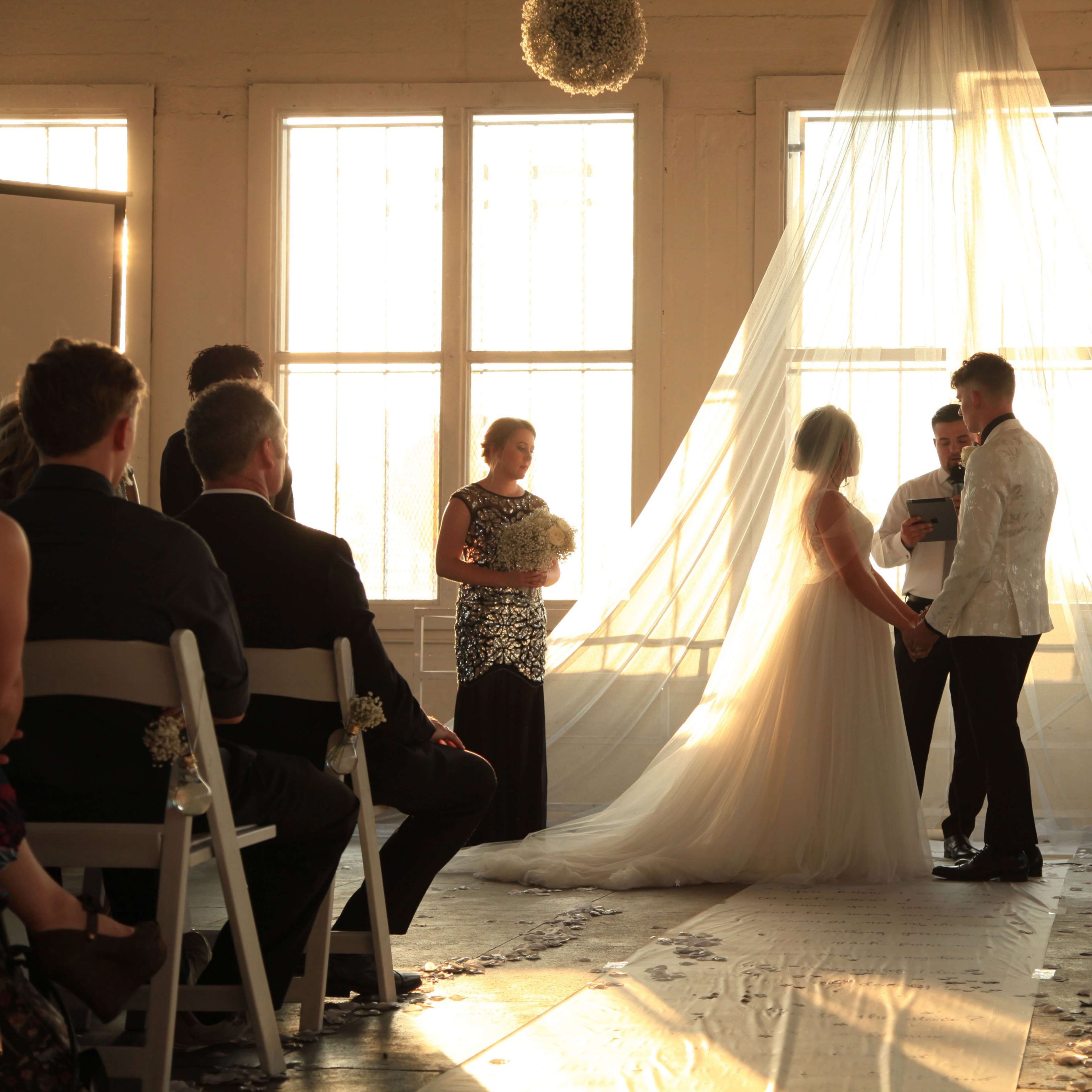 A couple stands in front of an altar while guests observe during a wedding ceremony in a sunlit room.