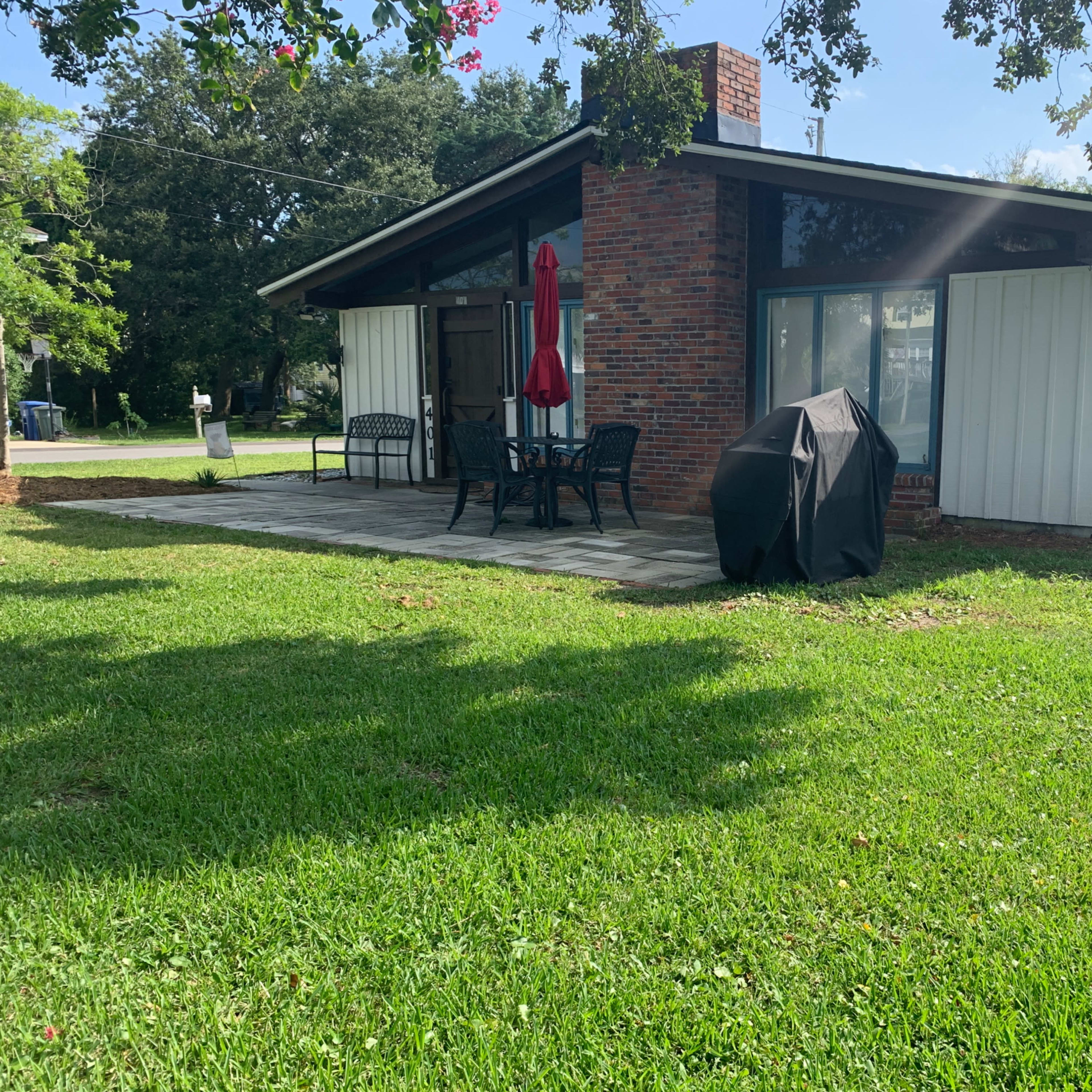 A single-story house with a red umbrella over a patio table, a charcoal grill, and grass surrounding it.