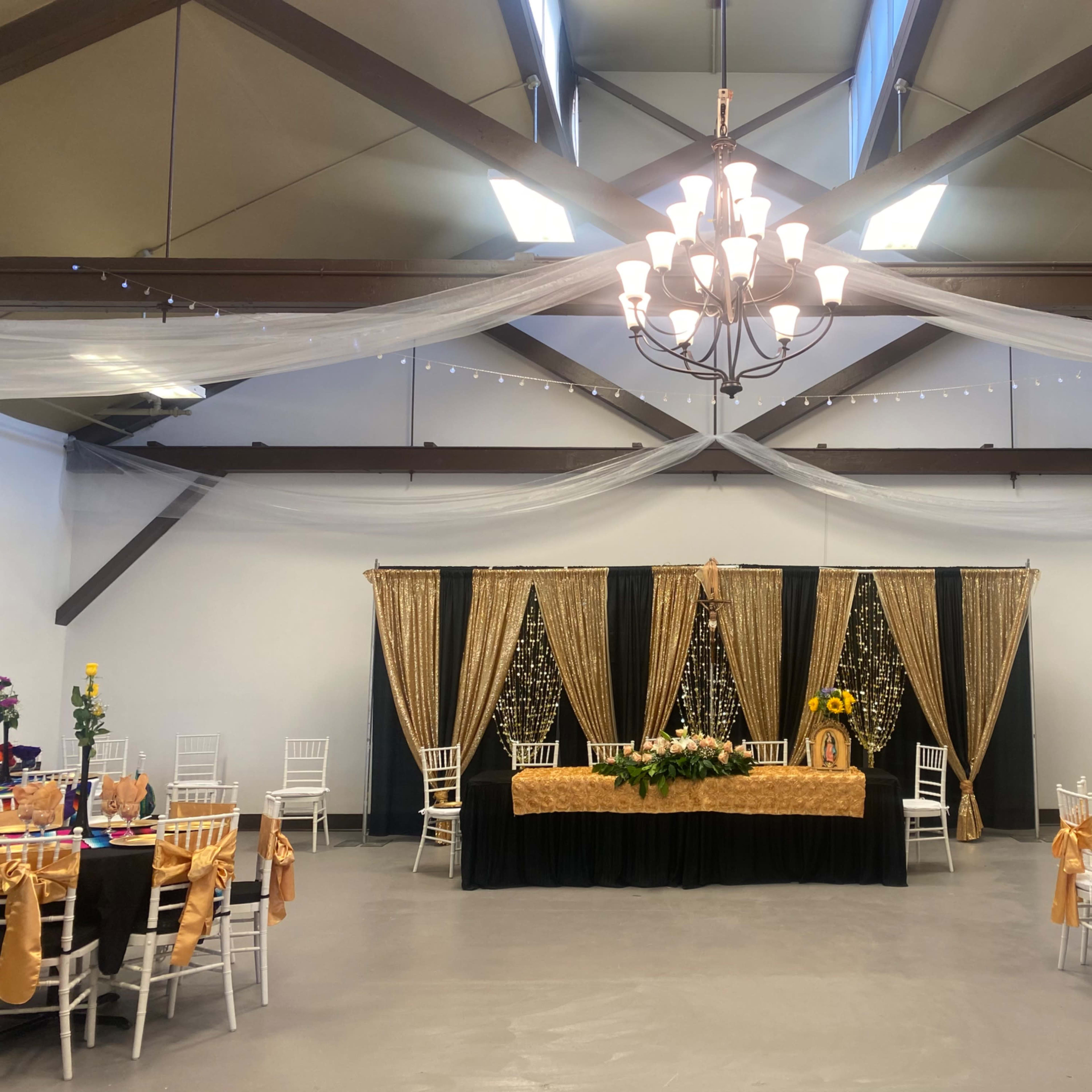 A spacious event hall features elegantly arranged tables with gold and white decorations, a large black table at the front, and draped fabric from the ceiling.