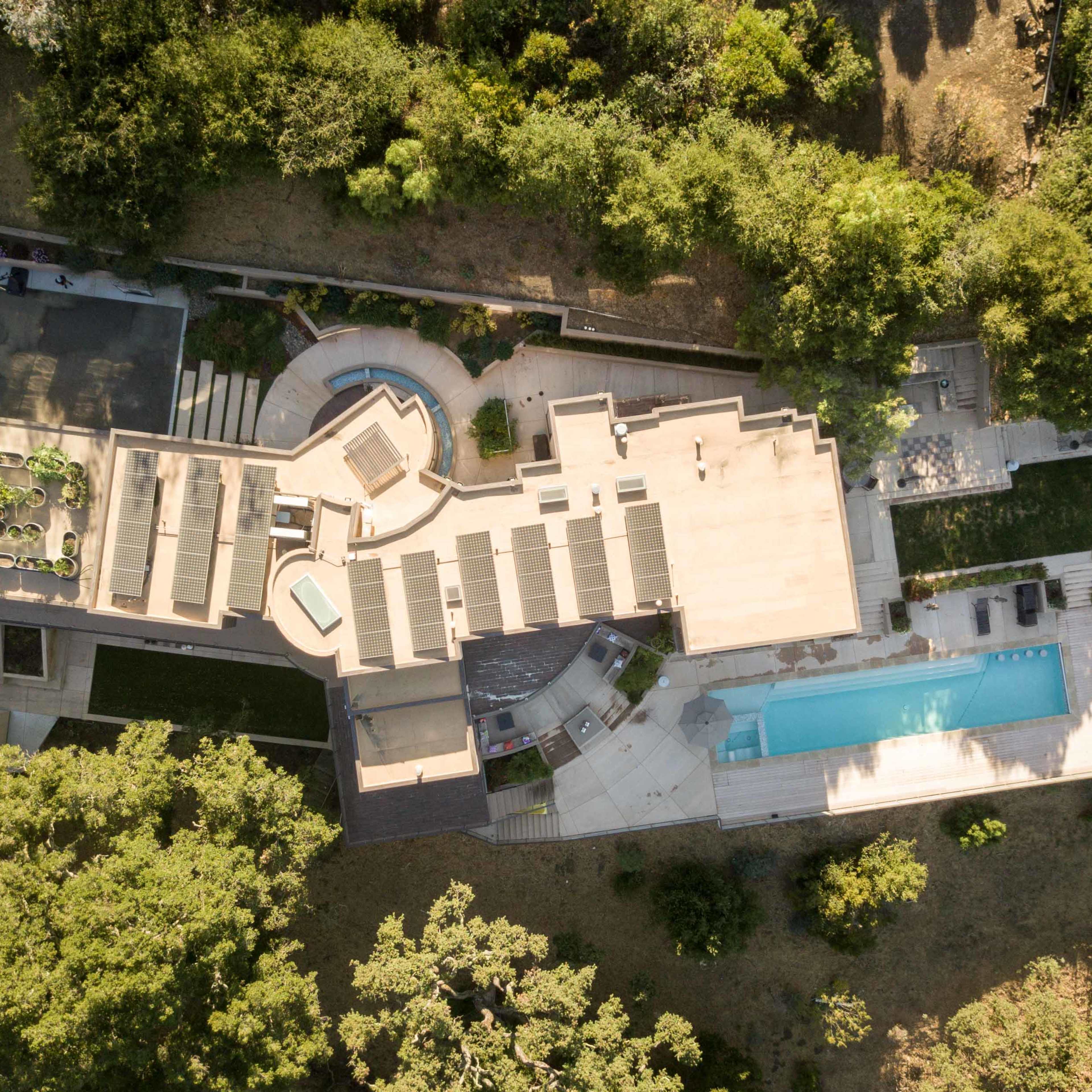 An aerial view showcases a modern house with a swimming pool, outdoor lounge areas, and landscaped gardens surrounded by trees.