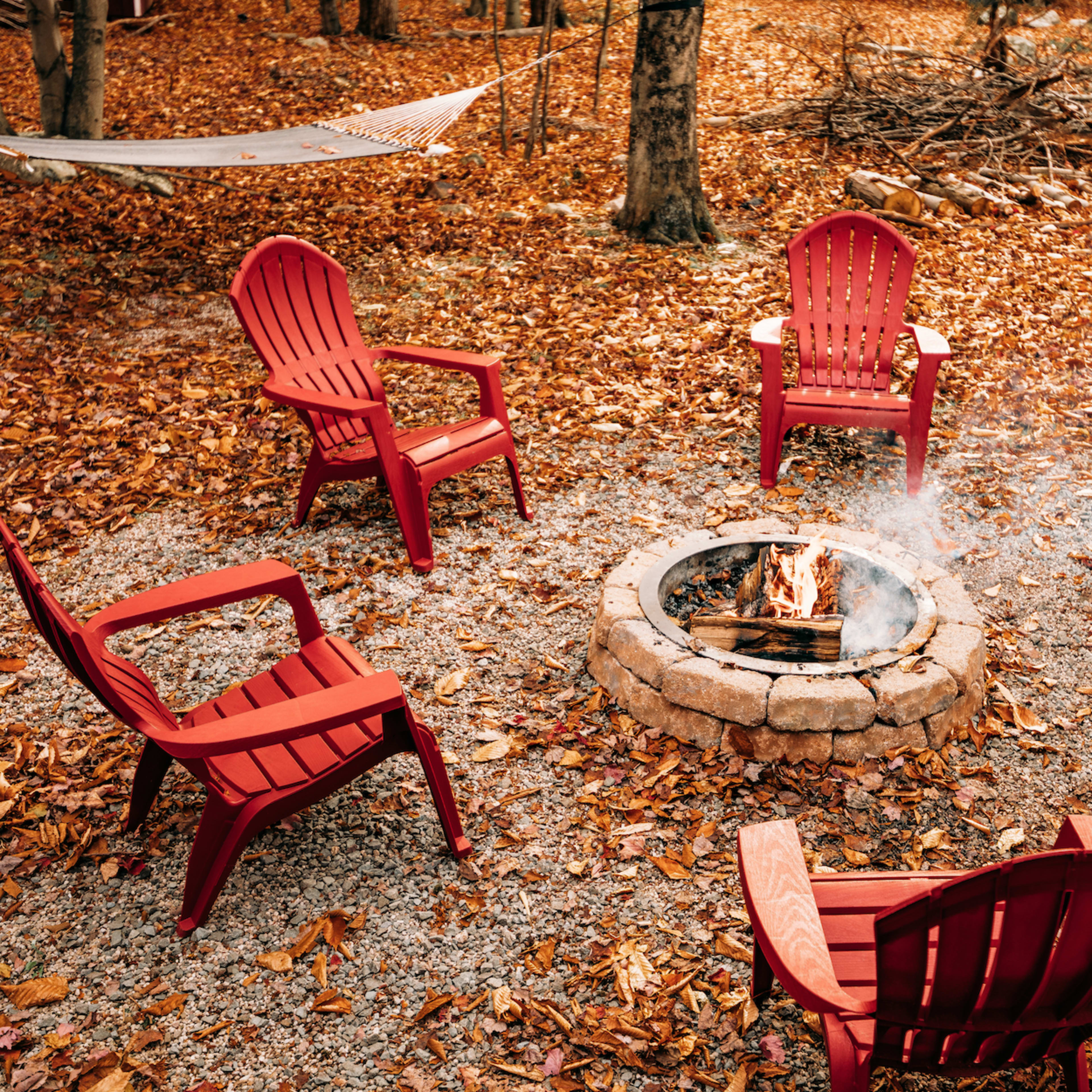 A circle of colorful chairs surrounds a stone fire pit with a small fire, set on a ground covered in fallen leaves.