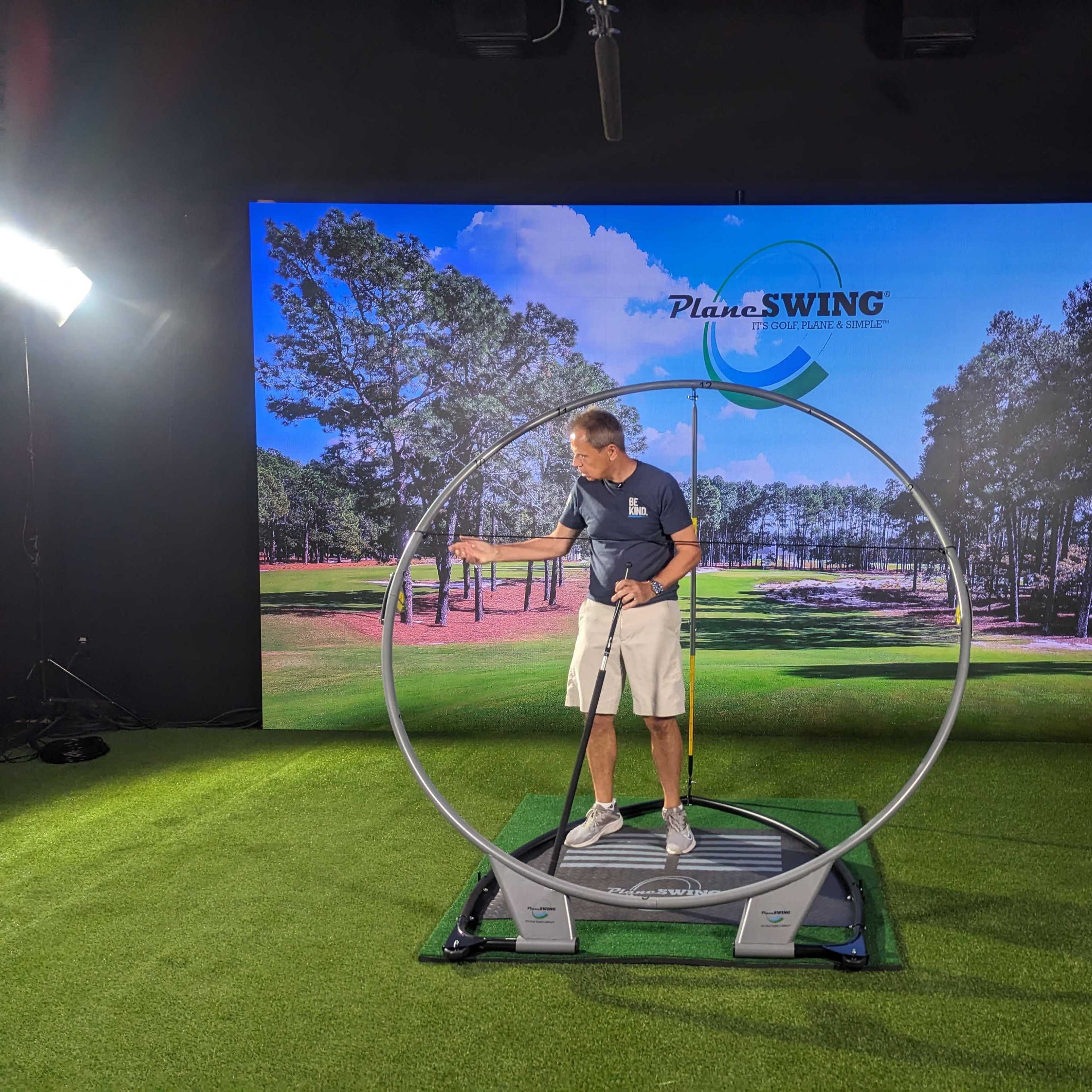 A golfer is demonstrating a swing technique using a training device in a golf simulation area with a backdrop of a golf course.