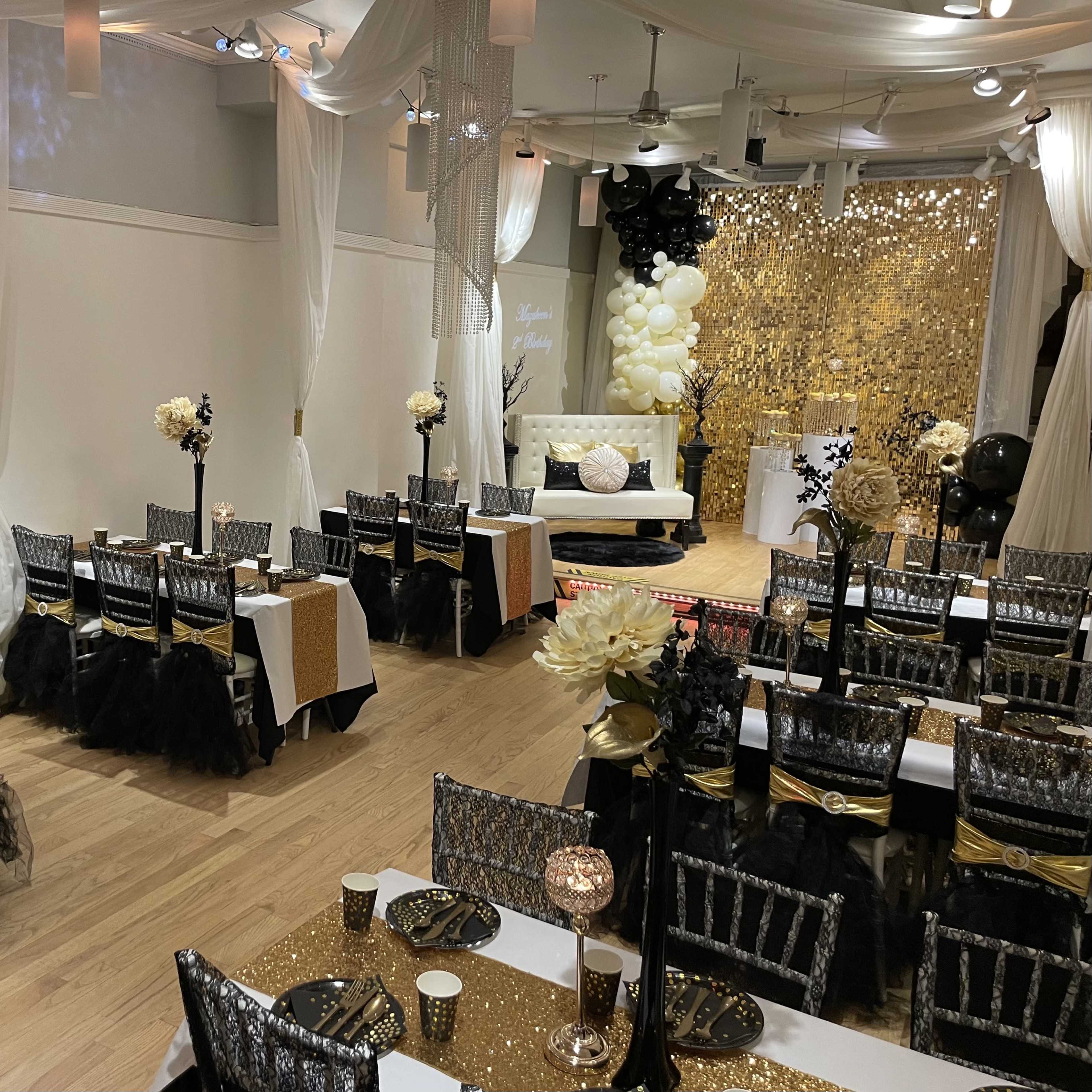 An elegantly decorated event space featuring tables set with white and gold tablecloths, a backdrop of golden sequins, and floral centerpieces.