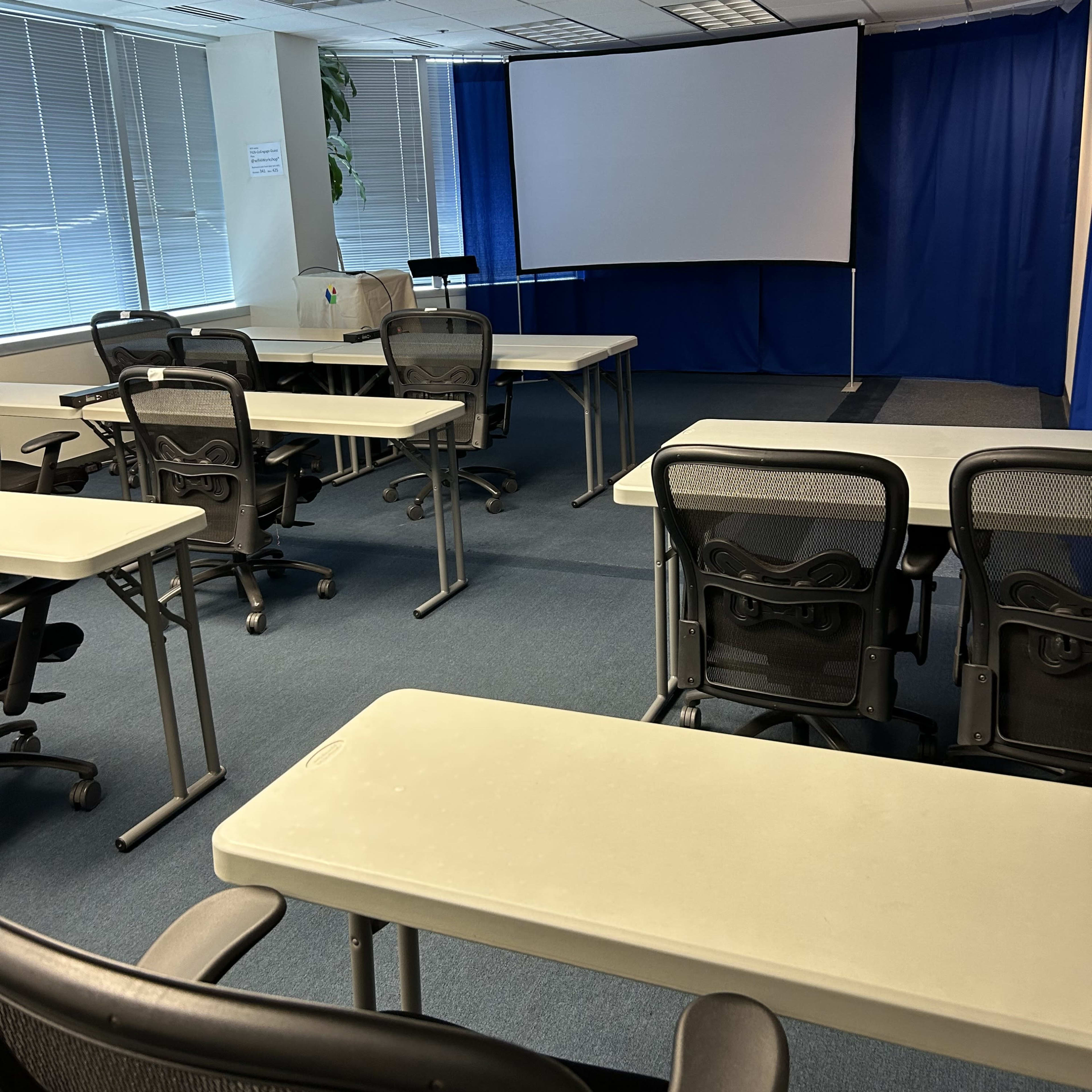 An empty classroom with several tables and chairs arranged facing a large projection screen at the front.