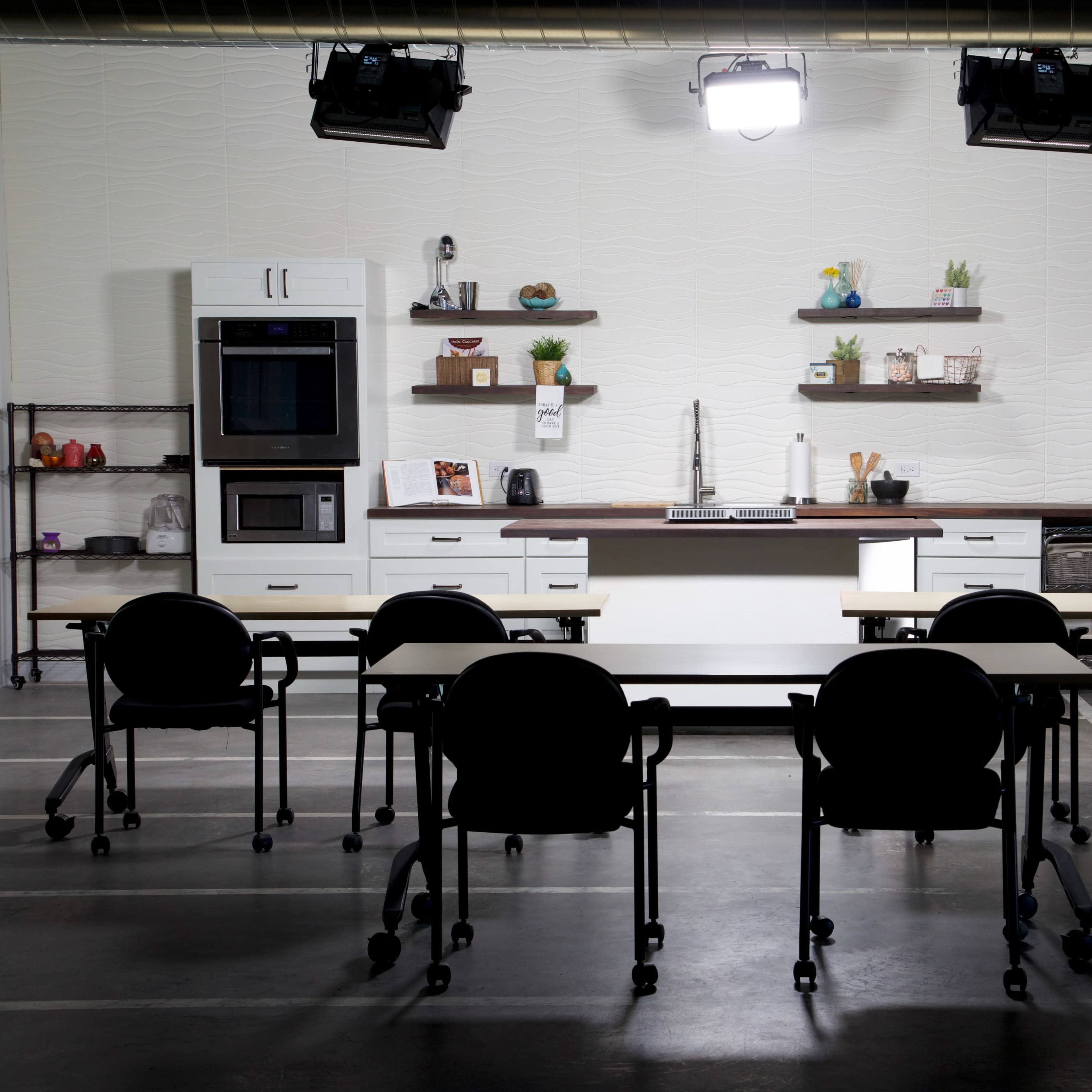 A modern kitchen setup features a stainless steel refrigerator, an oven, and minimalist shelving, with six black rolling chairs positioned around two tables.