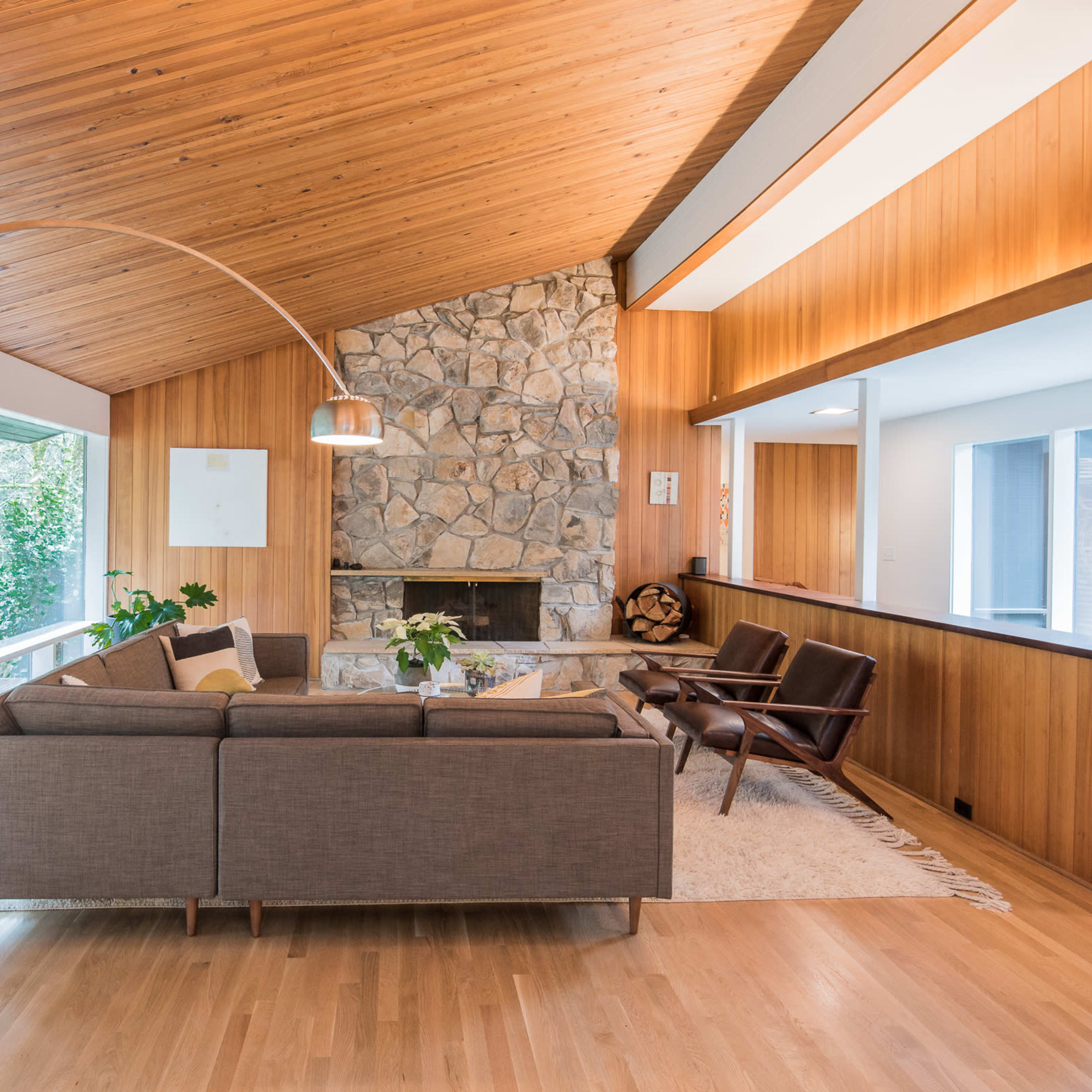 The living room features a stone fireplace, a curved floor lamp, and large windows that offer views of the surrounding greenery.