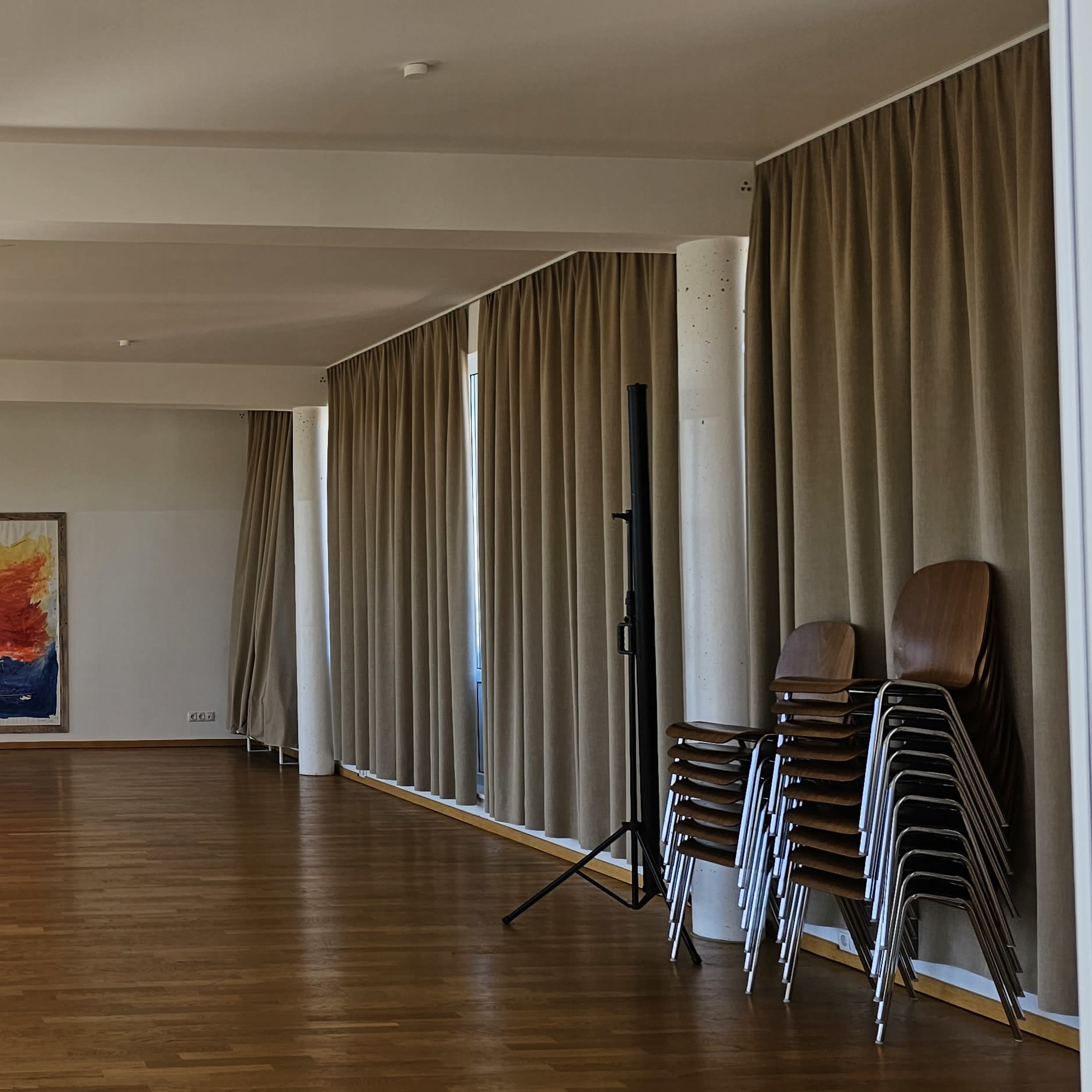 A spacious interior room with wooden flooring, large windows, and a colorful painting on the wall, alongside stacked chairs and curtains.