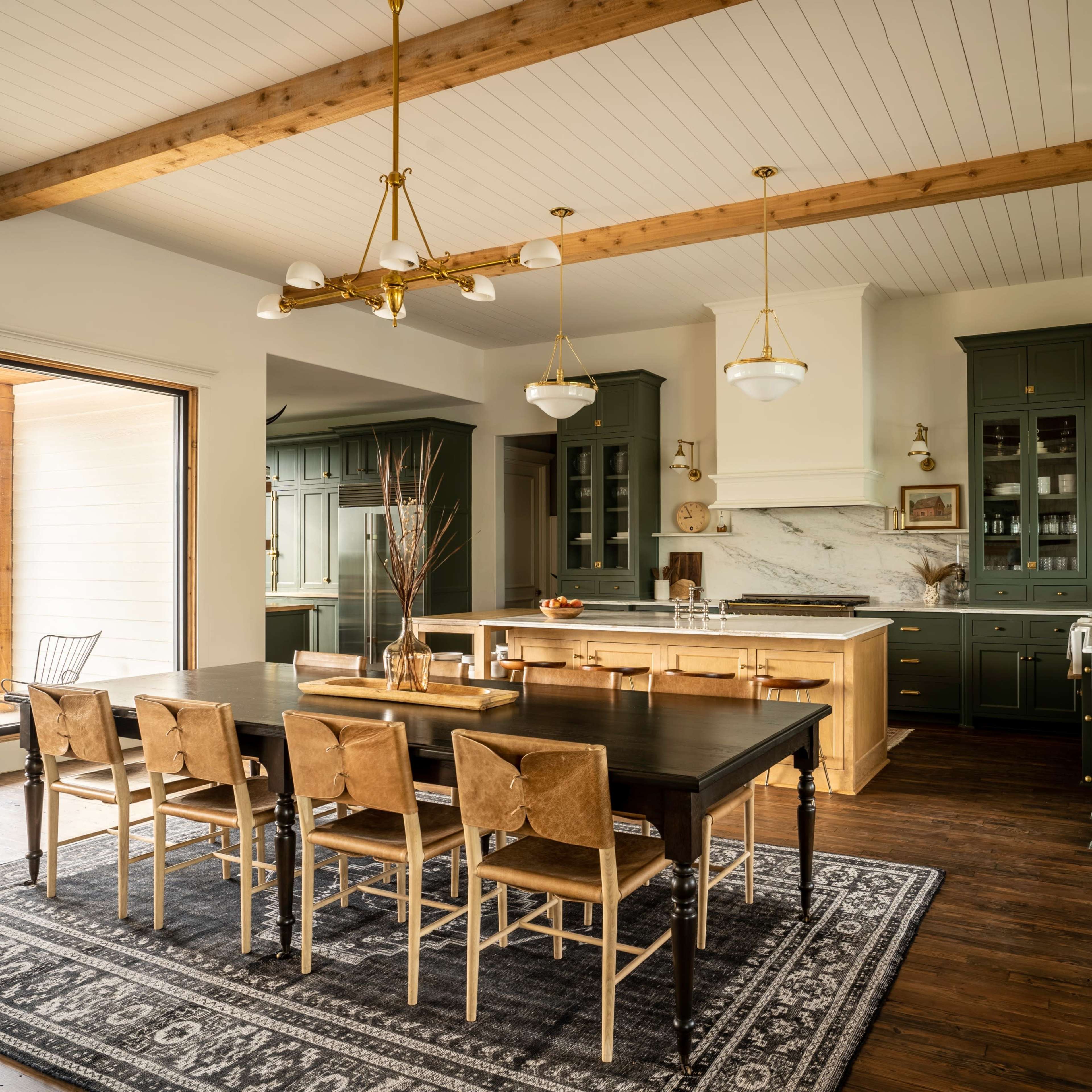 A spacious kitchen with green cabinetry, a large wooden dining table surrounded by chairs, and large windows that offer views of the outdoors.