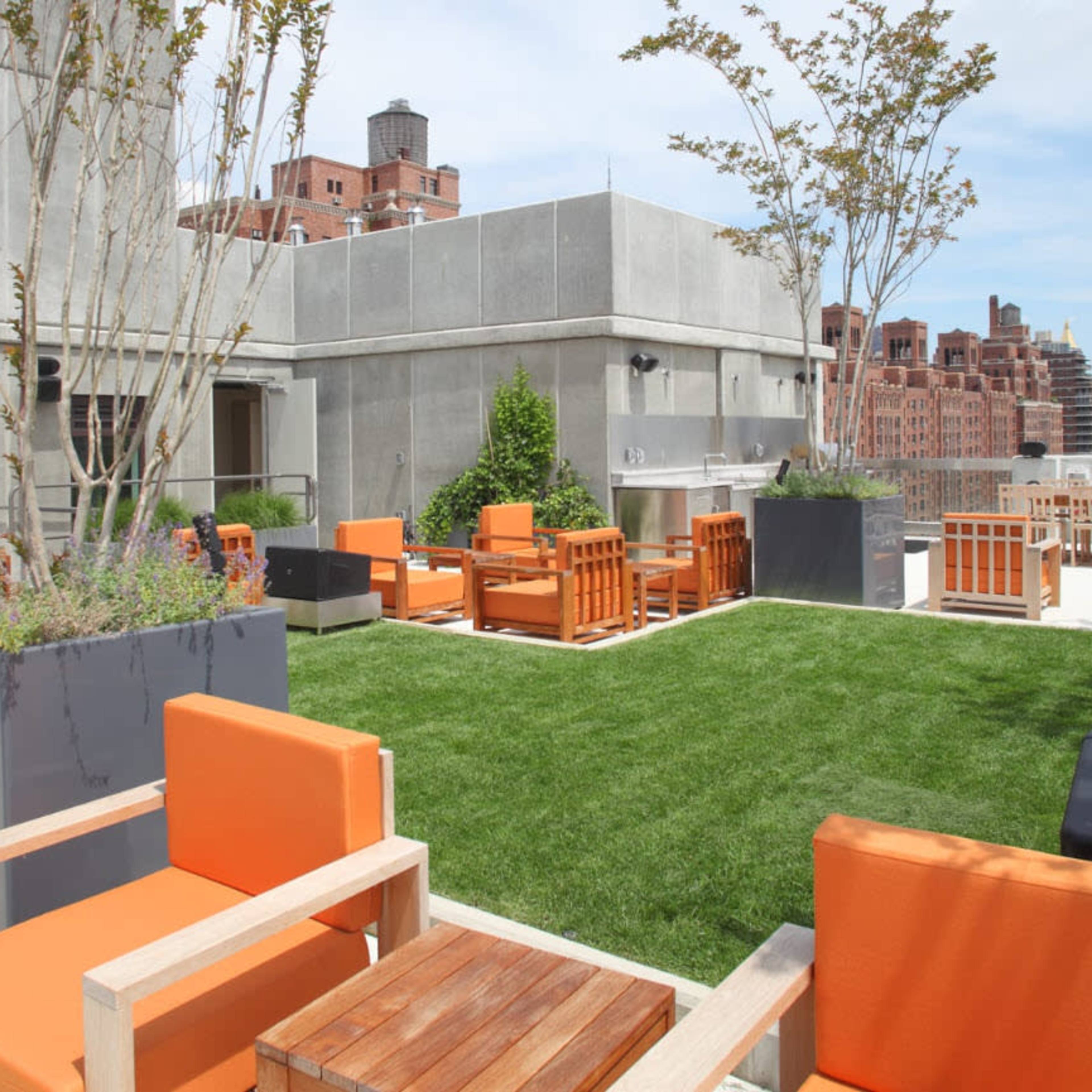 A rooftop patio featuring orange chairs, wooden tables, and potted trees, overlooking a city skyline.