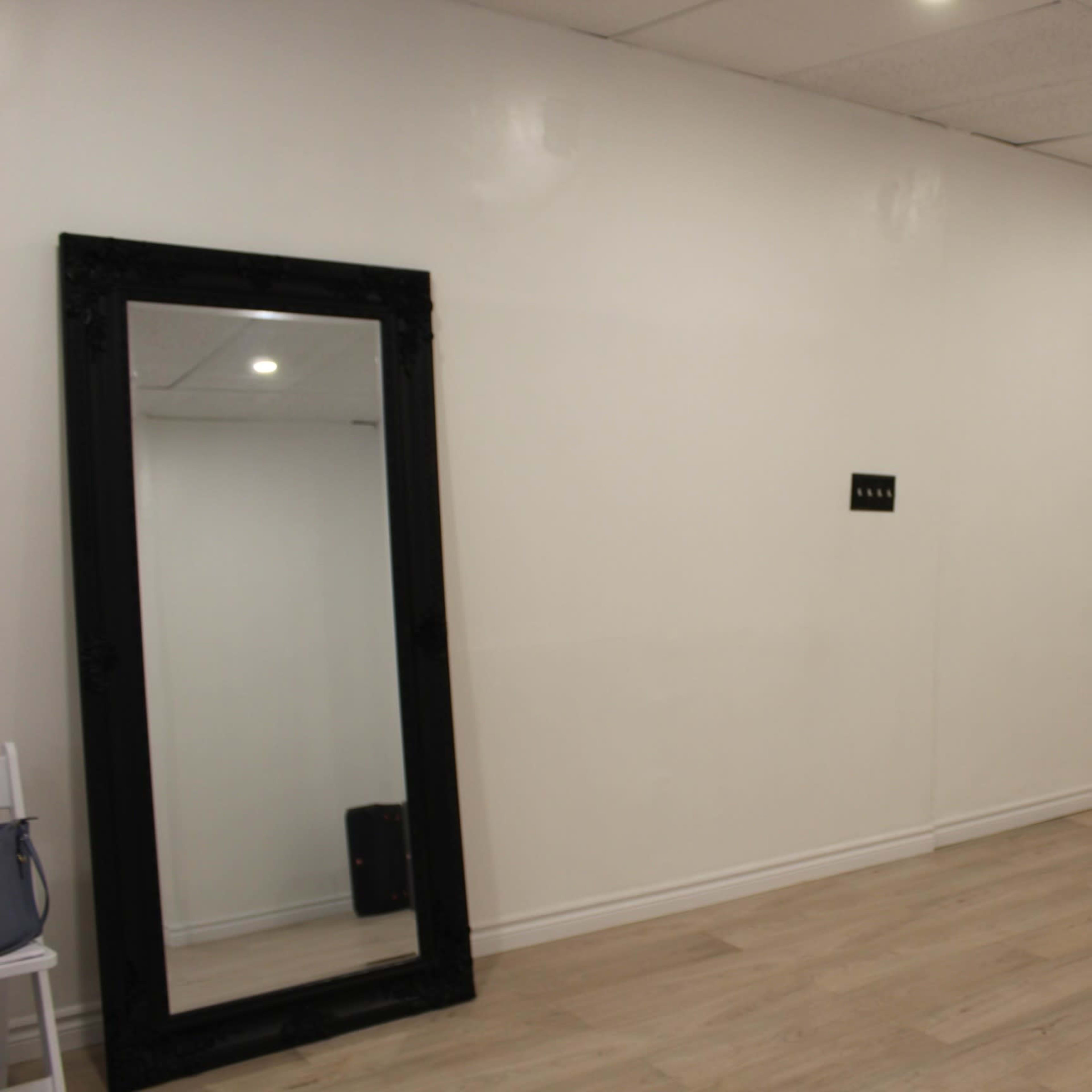 A spacious, empty room with light wooden flooring, white walls, and a large black-framed mirror on one side.