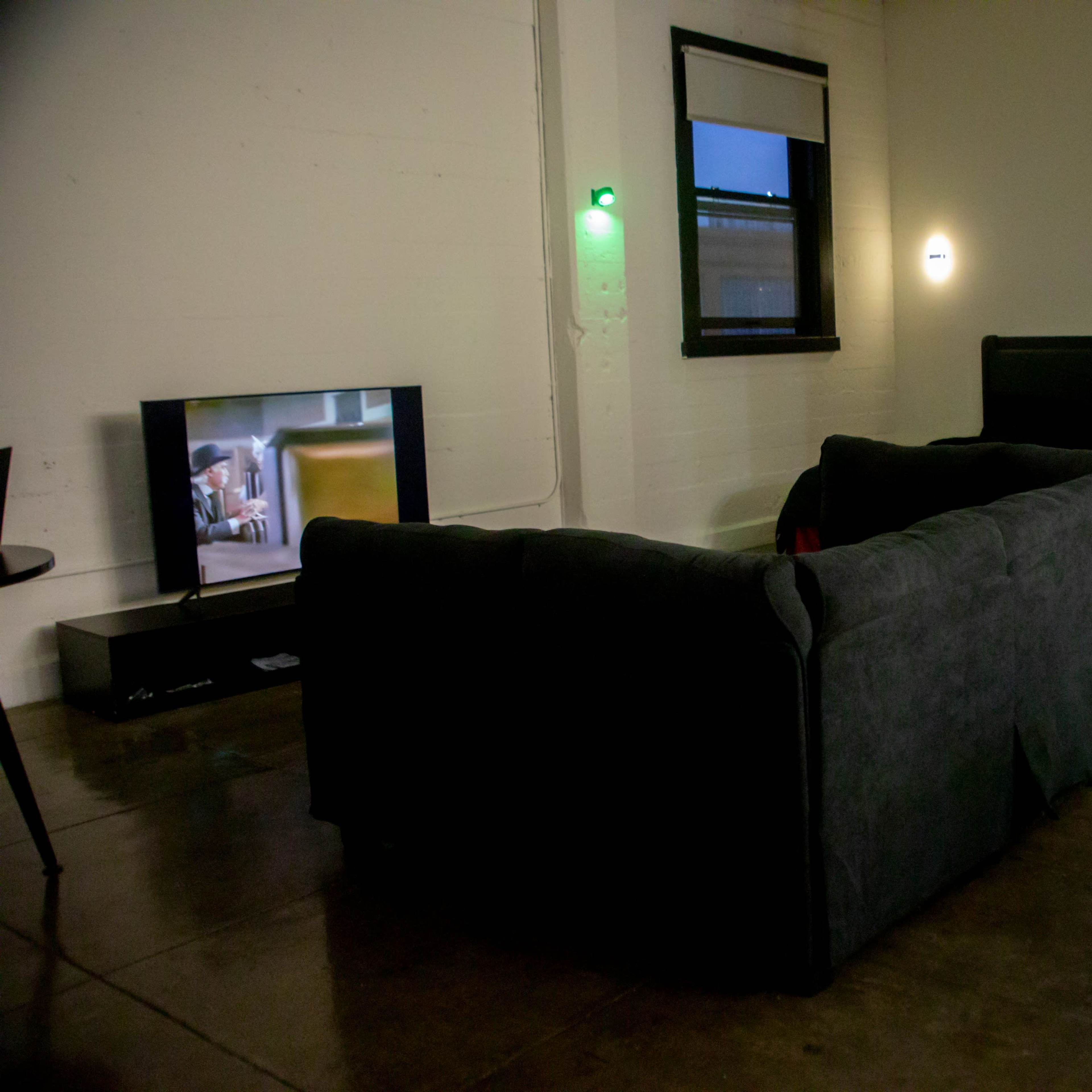 A living room features a gray sectional couch facing a television, with a door and windows in the background.