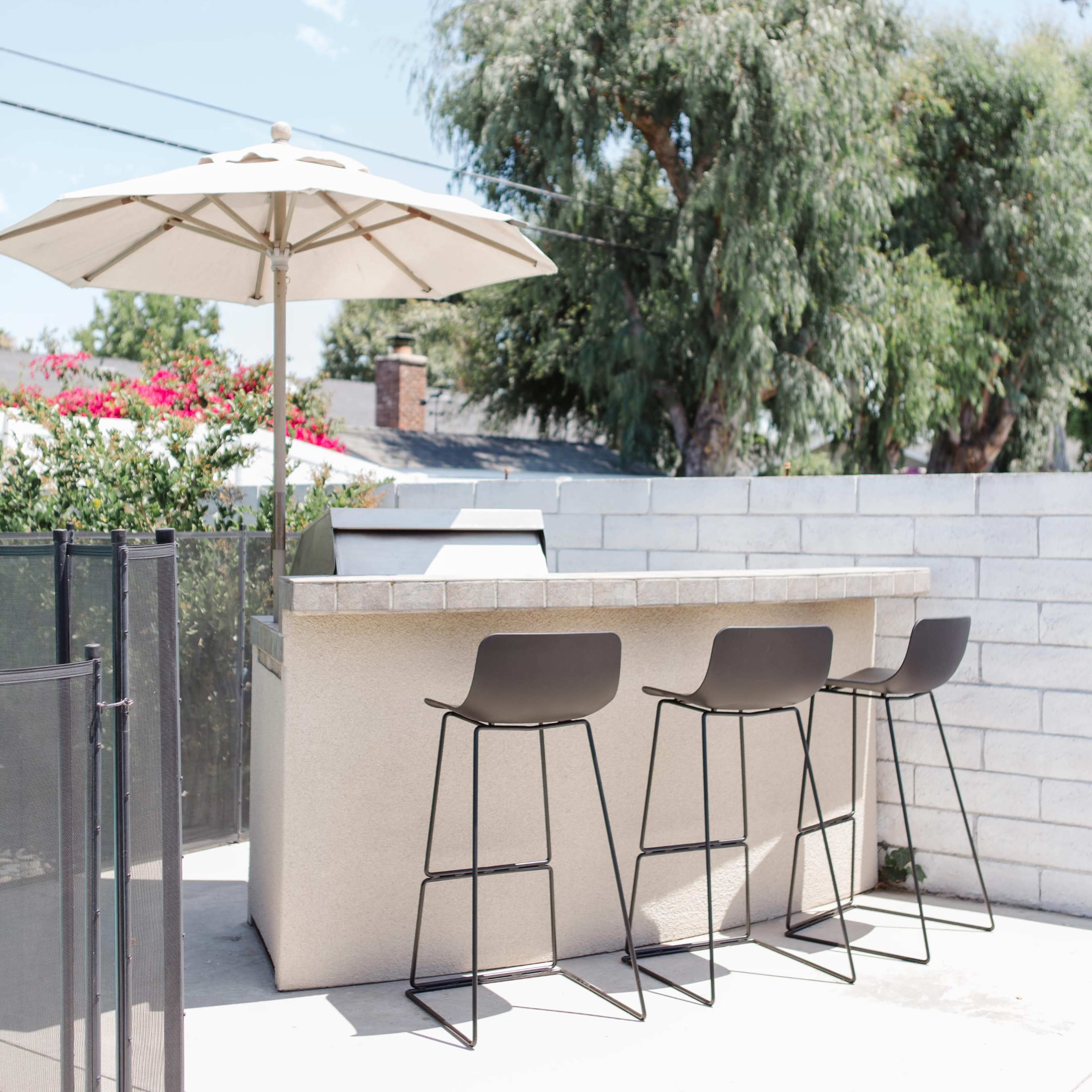 An outdoor bar area with three black stools, a grill, and an umbrella, surrounded by greenery and a concrete wall.