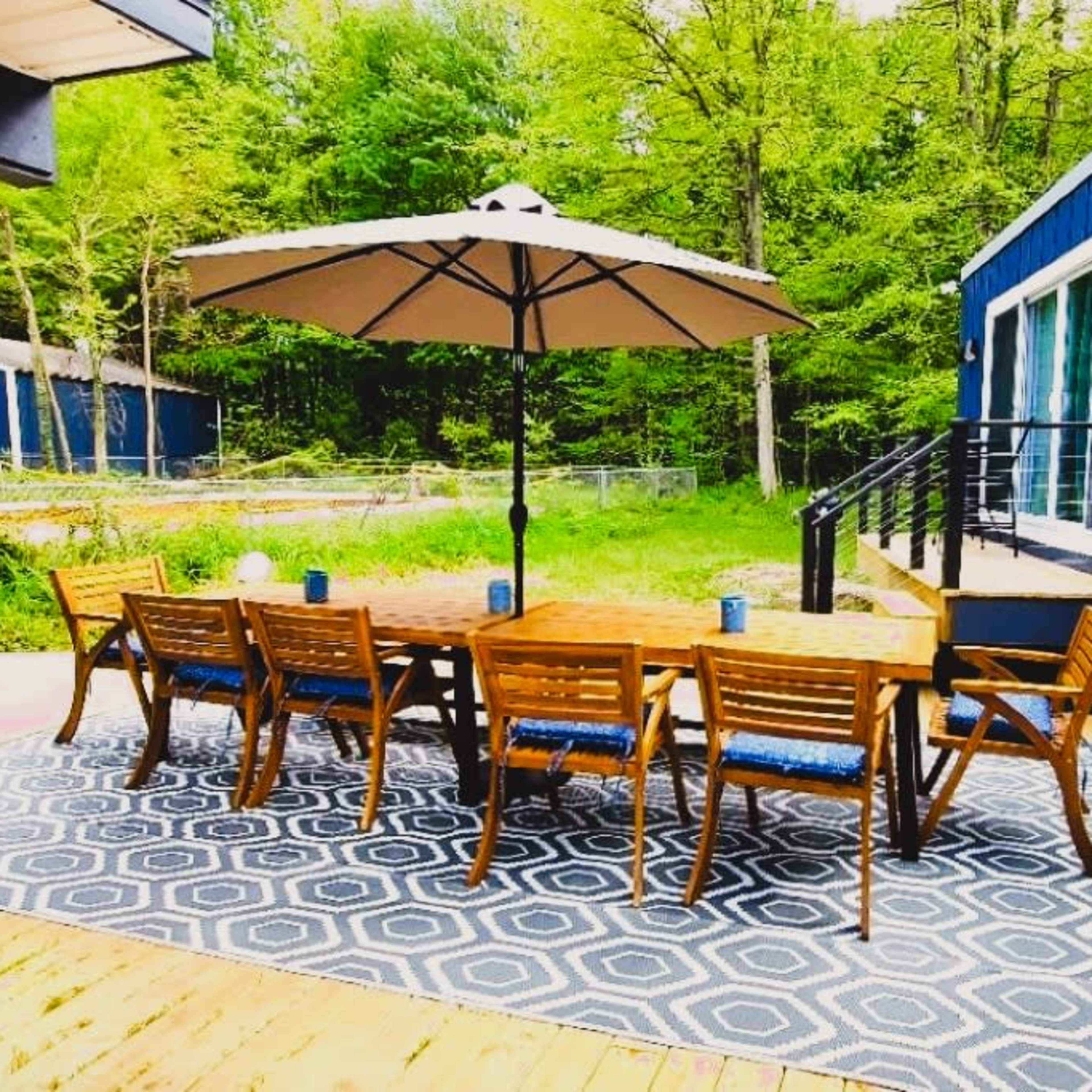 A large wooden table with chairs is set under an umbrella on a deck surrounded by green trees.